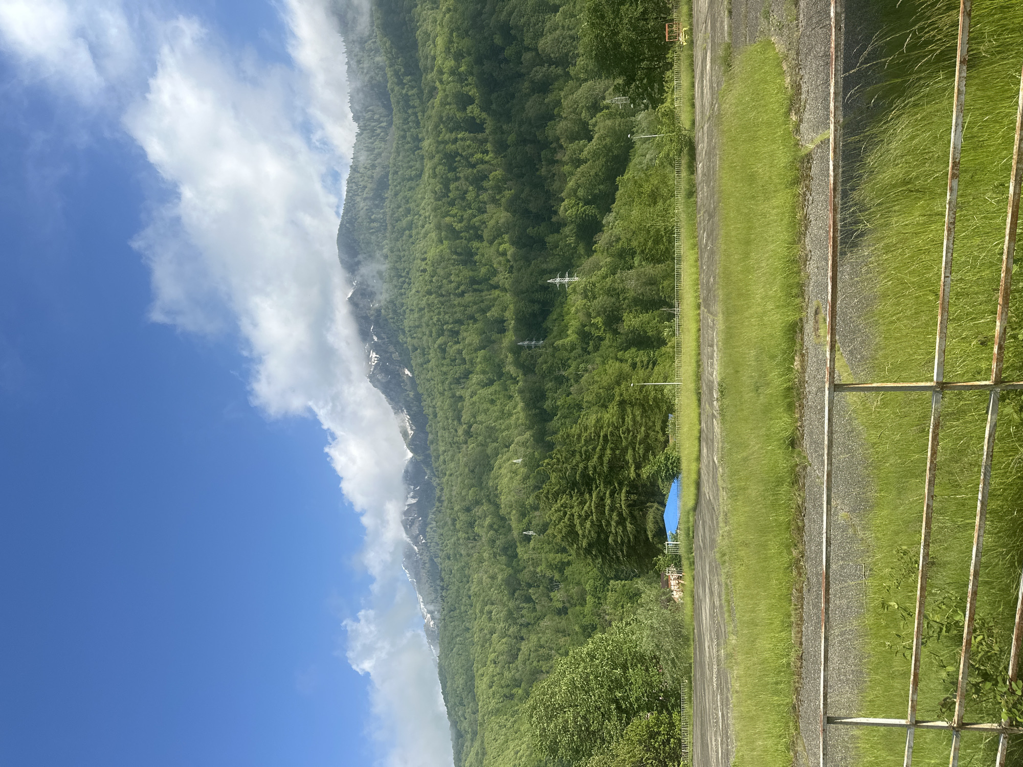 The image depicts a landscape in Krasnaya Polyana, Russia. A mountain, partially obscured by clouds, is covered in dense green forest. A few patches of snow are visible along the mountain's slope. The foreground features a grassy area alongside a paved surface with a metal fence. In the middle ground, a blue canopy is situated amongst the trees. Power lines can be seen running through the forested area. The sky is blue with white clouds.