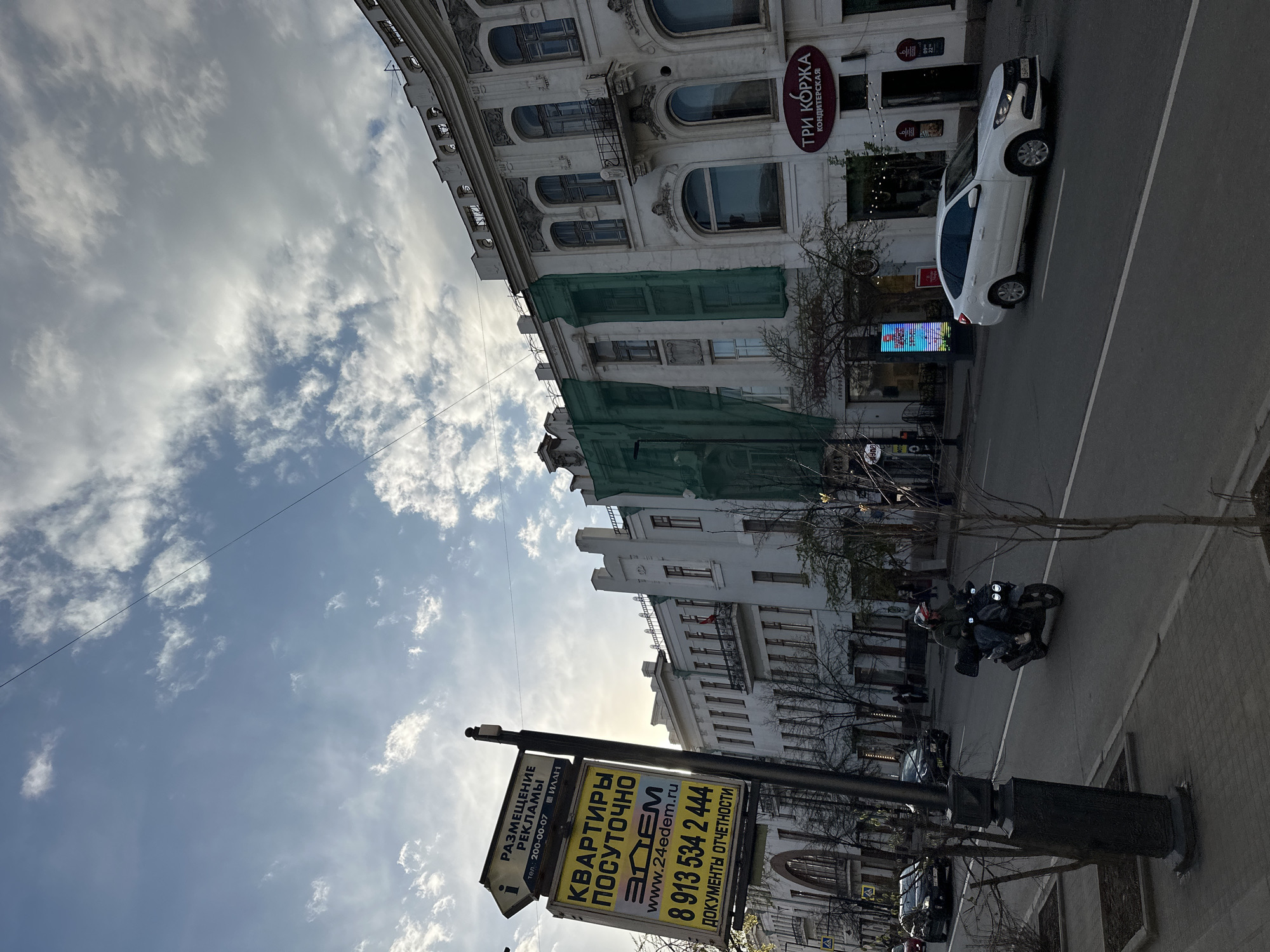 The image captures a street scene in Krasnoyarsk, Russia. The foreground features a street sign advertising apartment rentals by the day. The signage includes the business name "Edem" and a phone number. A building under construction is partially obscured by green scaffolding. A white car is parked along the street, and a motorcycle with two people is traveling on the street. A shop named "Три Коржа" (Three Korzha) is visible in the background. The sky is partially cloudy.