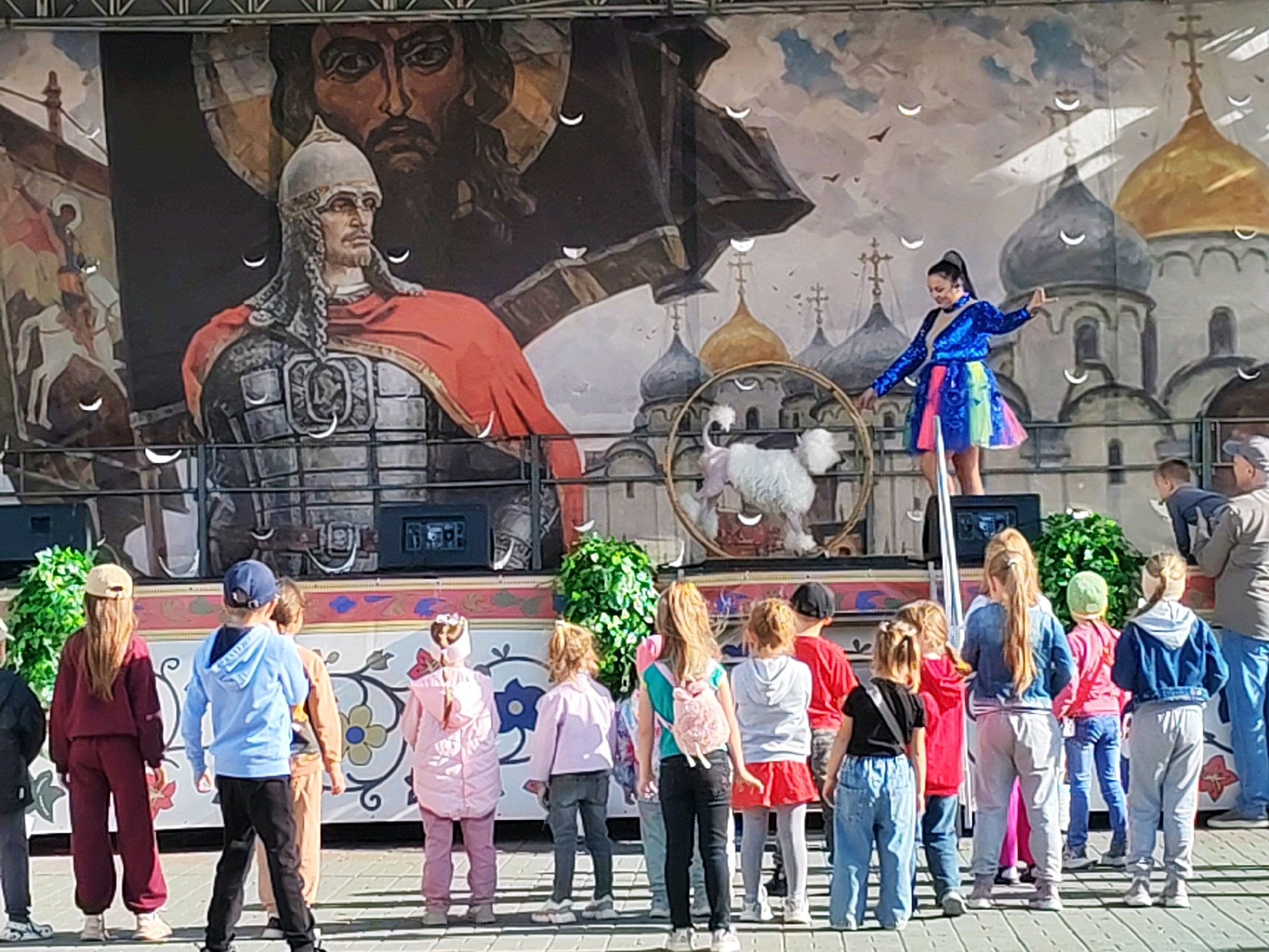 An outdoor daytime event is underway in front of a large stage backdrop that depicts a historical mural. The mural features a stern-faced warrior in armor and a red cloak, with a larger, dark-robed figure behind him. In the middle ground of the mural are stylized depictions of domed buildings, possibly churches. On the stage, a woman in a sparkling blue jacket and a colorful, iridescent skirt is performing. She is standing on a raised platform and appears to be interacting with a white, fluffy prop animal, possibly a dog or a stuffed toy, which is inside a circular hoop. A group of children are gathered in front of the stage, watching the performance. They are of various ages and are dressed casually. Some are wearing hats and hoodies. To the left of the stage, there are speakers and some greenery. The ground in front of the stage is paved with light-colored tiles. The overall scene suggests a public performance or festival.