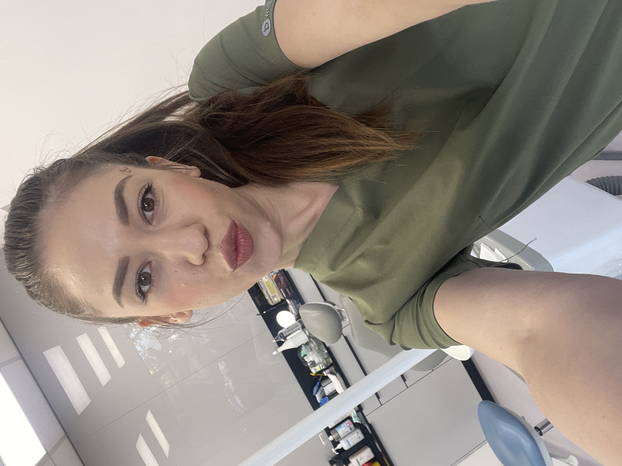 A woman is taking a selfie. She has brown hair, fair skin, and is wearing a green top. She is inside what appears to be a dental office, given the dental equipment in the background. The exact location in Sofia, Bulgaria, cannot be determined from the image.