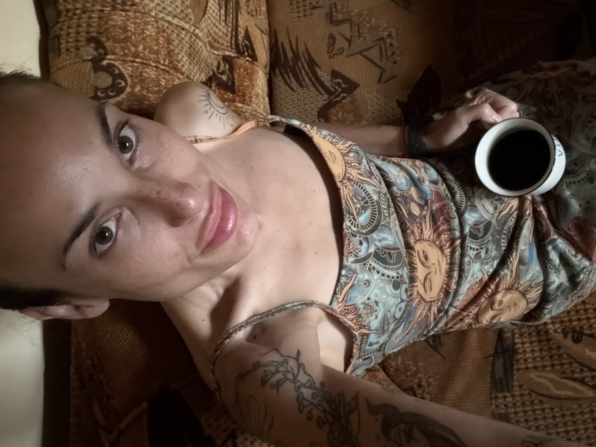 A woman with tattoos on her arms is sitting on a couch, holding a white mug of coffee. She is wearing a patterned dress with sun motifs. The couch appears to be upholstered in a brown, textured fabric. The lighting suggests an indoor setting. The image is a close-up, focusing on the woman's face and upper body. No specific location in Pleven, Bulgaria is identifiable from the visual information provided.