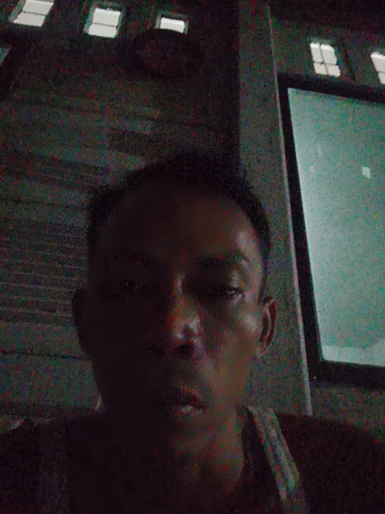 A low-resolution image depicting a person taking a selfie in a dimly lit room. The person has dark hair, and their face is partially obscured by shadow. Behind the person, there is a window with white panes, and a framed object. The location is possibly Bukit Ketok, Indonesia.