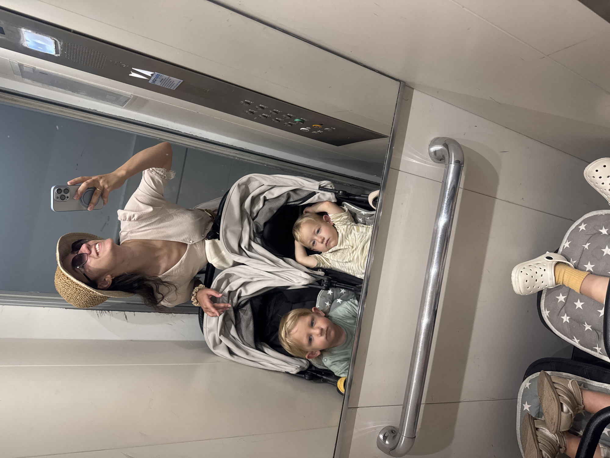 A woman wearing a straw hat and sunglasses is taking a selfie in a mirror inside what appears to be an elevator. Two children are in a double stroller facing the mirror. The elevator's control panel is visible above the woman in the mirror. The stroller and woman's reflection indicates the location is in Bat Yam, Israel. The interior is utilitarian, with a silver handrail visible on the wall.