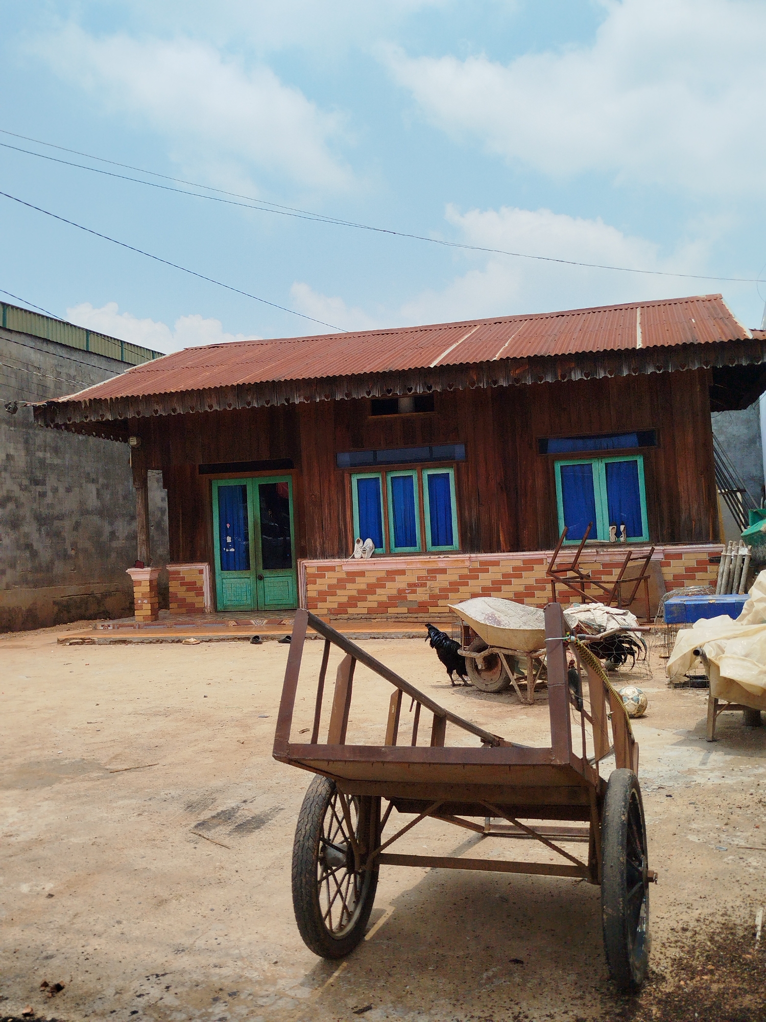 This daytime image depicts a rural or semi-rural scene in Thôn 5, Vietnam, under a bright blue sky with scattered white clouds. The central subject is a traditional wooden house with a corrugated reddish-brown metal roof. The house features vibrant turquoise-painted window frames and a central double door, with dark blue curtains visible behind the windows. Its base is constructed from bricks and tiles. Various items of footwear are placed outside the house, near the main door and one of the windows.
In the immediate foreground, a large, empty, two-wheeled brown handcart stands on a dusty dirt ground. To the left of the house, a dark-feathered rooster is seen near a white wheelbarrow. Another chicken is partially visible further right, close to a soccer ball and objects covered by a beige tarp. A textured concrete wall runs along the left side of the scene, and utility lines are visible crossing the sky. No human activity is apparent, presenting a static, tranquil village scene. There is no visible text in the image.
