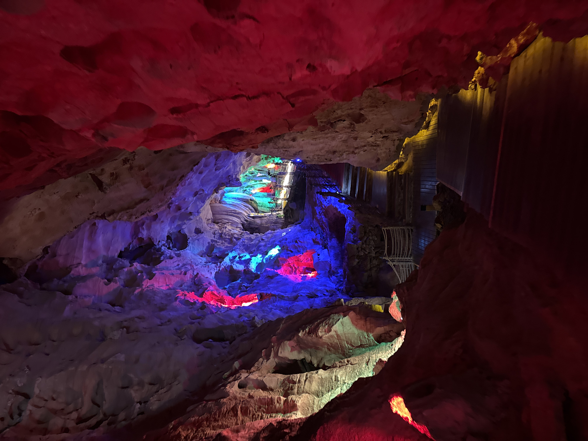 The image shows the Leigu Cave system in Zhuoshui, China. The cave interior features rock formations illuminated with red, blue, and green lights. A wooden walkway is built along the cave wall.