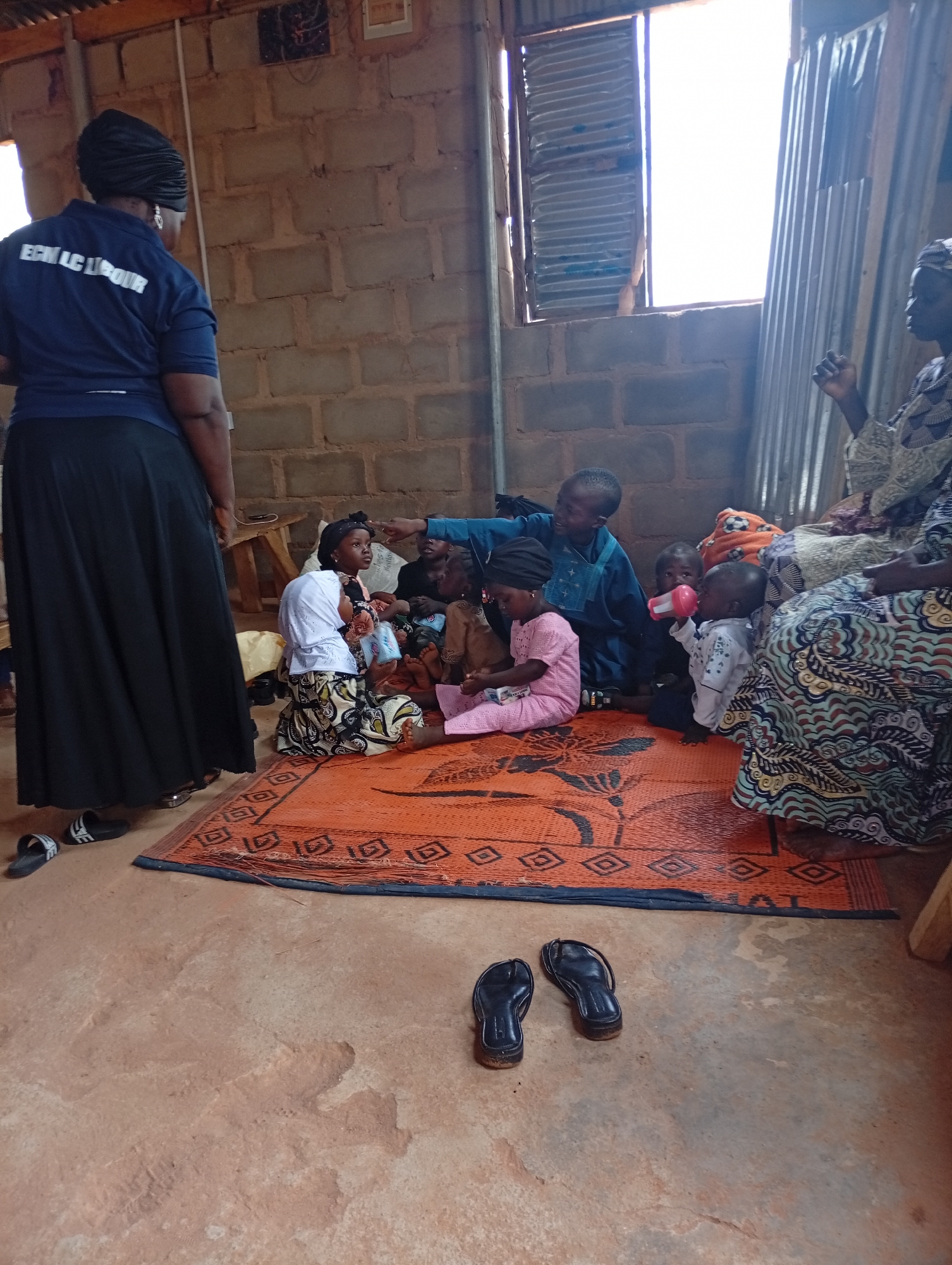 An indoor scene depicts a group of people, primarily women and children, gathered in a room with a dirt floor and brick walls. The room is sparsely furnished. On the left, a woman wearing a blue shirt with white lettering and a black headscarf stands facing away from the camera, her long black skirt pooling around her. In the center, several children are seated on an orange mat. One child, wearing a white headscarf and patterned skirt, sits closest to the camera and appears to be looking to the right. Next to her, a child in a pink outfit looks to the left. Another young child in a white shirt drinks from a pink bottle. Behind the children, a person in a blue garment is partially visible, leaning forward. On the right side of the frame, an adult, likely a woman, is seated, wearing a colorful patterned dress. The background features a rough brick wall, a window with corrugated metal blinds, and a wooden stool. Black flip-flops are visible on the floor in the foreground and near the standing woman. The overall atmosphere suggests a community gathering or educational session.