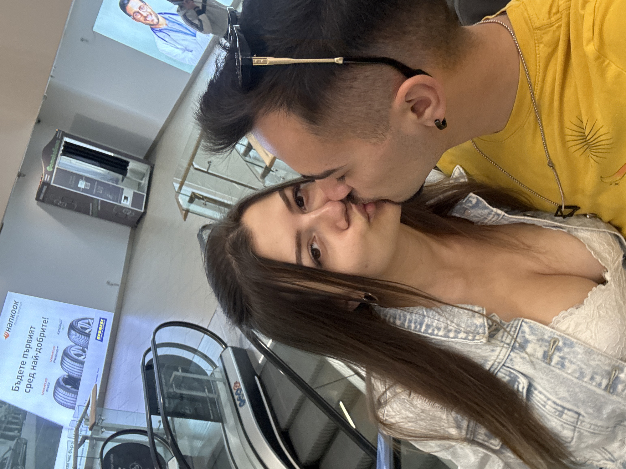 A photo depicts a couple sharing a kiss. The woman has long brown hair, a white lace top, and a light blue denim jacket. The man has dark hair, a black earring, sunglasses on his head, a yellow t-shirt, and a necklace with a pendant. They appear to be inside a commercial building, with an escalator in the background. There is a poster with the Hankook logo and Bulgarian text, suggesting they may be in Sofia, Bulgaria.