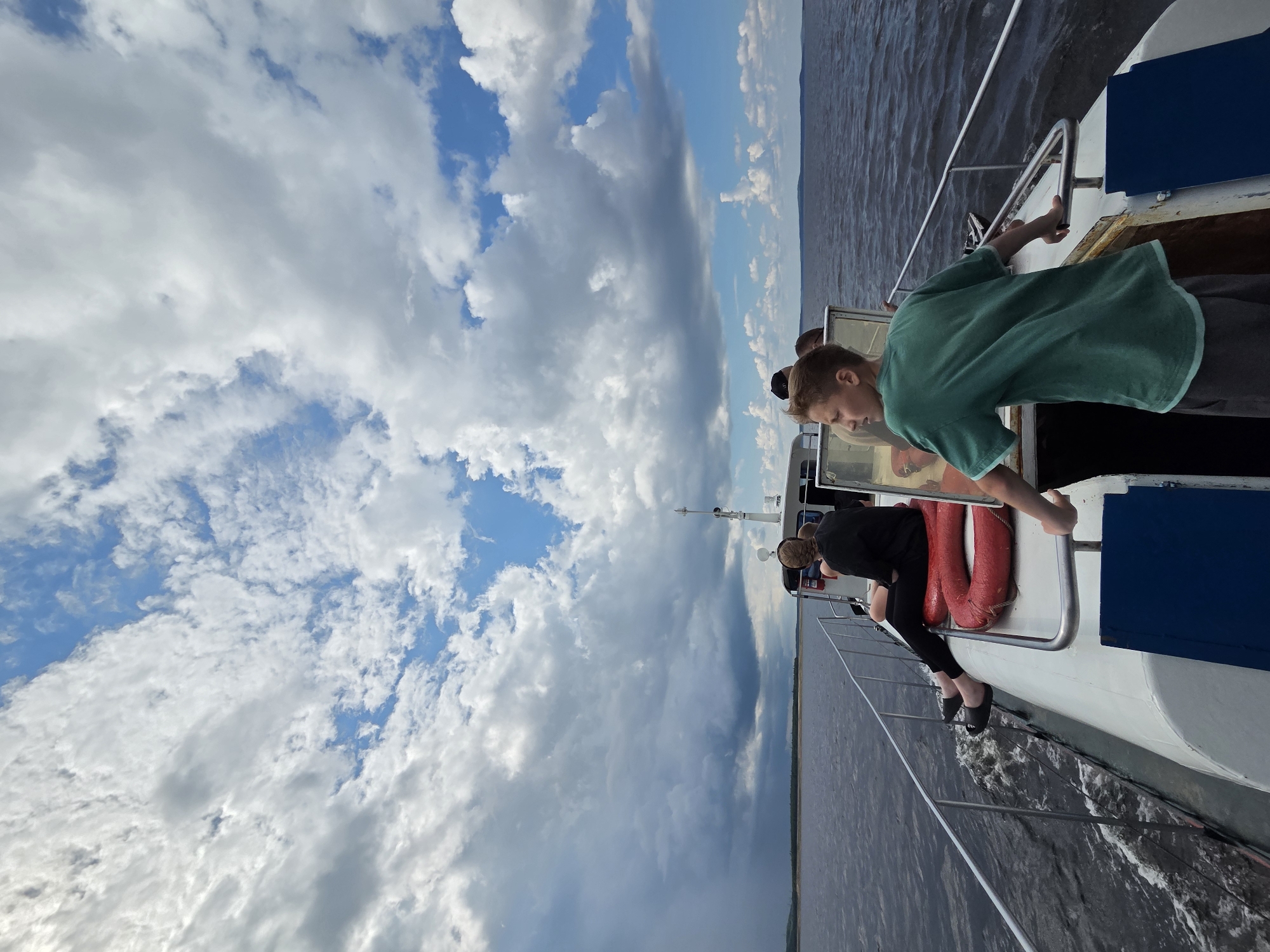 On a boat navigating calm waters under a partly cloudy sky, a young boy in a green t-shirt and gray shorts stands by the railing, his hands on the metal bar as he looks out to the left. Behind him, other passengers are seated. To the left, a person in a black shirt and leggings sits on a life preserver, their legs extended. The boat travels across a body of water with a distant, flat shoreline visible. The sky above is filled with dramatic, white and gray clouds. The location appears to be the city of Talakan, Russia.