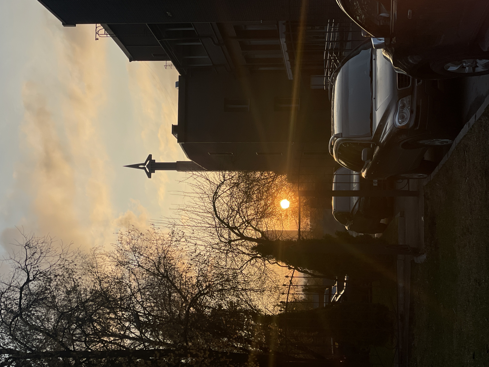 The image depicts an outdoor urban or suburban scene captured at sunset. The photograph is oriented vertically, rotated 90 degrees counter-clockwise from its natural landscape view.
The central and most striking element is the setting sun, a brilliant circular disc positioned low on the horizon on the right side of the frame. Its intense light bathes the sky in warm orange and yellow hues, creating a strong lens flare and silhouetting much of the foreground and midground. The sky above the horizon features scattered, soft clouds, contributing to the warm color palette.
In the foreground and midground, numerous deciduous trees with bare branches stand out as dark, intricate silhouettes against the bright sky. On the left side of the image, the dark outlines of several multi-story buildings, likely residential or commercial, are visible. Further into the midground, to the left of the sun, a tall, distinctive spire or monument-like structure with a pointed top and a cross-like or star-like embellishment is silhouetted against the radiant sky.
On the right side of the frame, multiple cars are parked, with a dark SUV, possibly a Volvo XC90, prominently featured in the midground, facing the viewer. The direct sunlight catches its contours, revealing some metallic reflections and details. A patch of green grass is visible in the bottom right corner of the image.
No people are visible. The scene evokes a calm and peaceful feeling typical of dusk. There are no discernible text or unique architectural elements that clearly indicate a specific location.