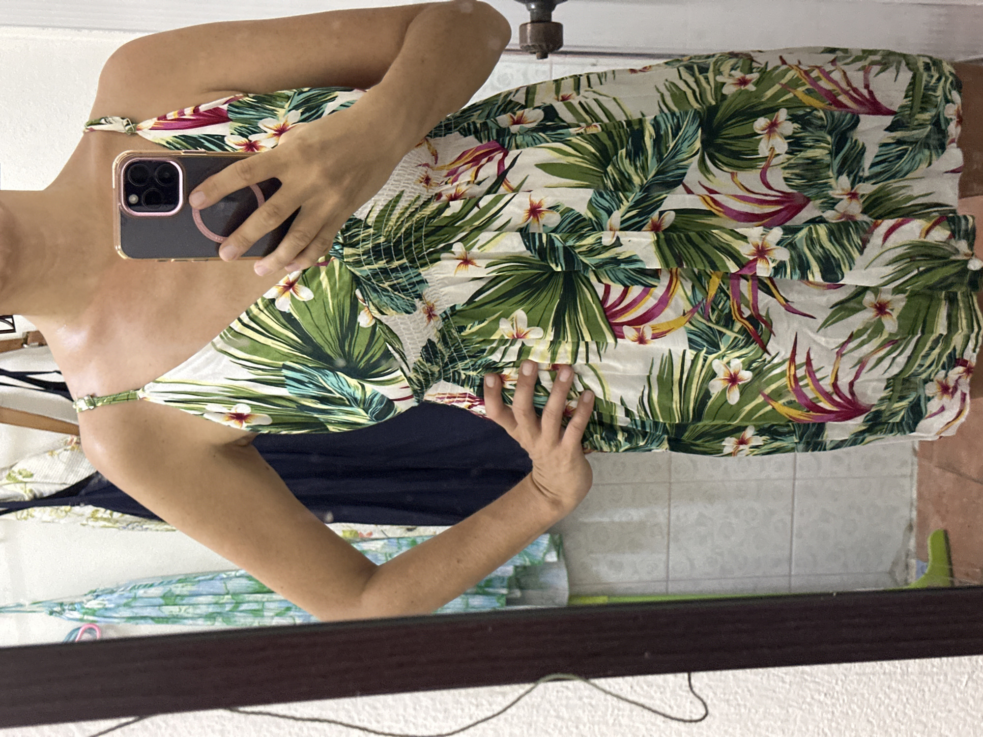 A woman is taking a selfie in a mirror, likely located in a residential setting in ตำบลเกาะพะงัน, Thailand. She is wearing a dress with a tropical floral pattern and holding a black smartphone. The background shows a wardrobe with other clothes. The floor is tiled.