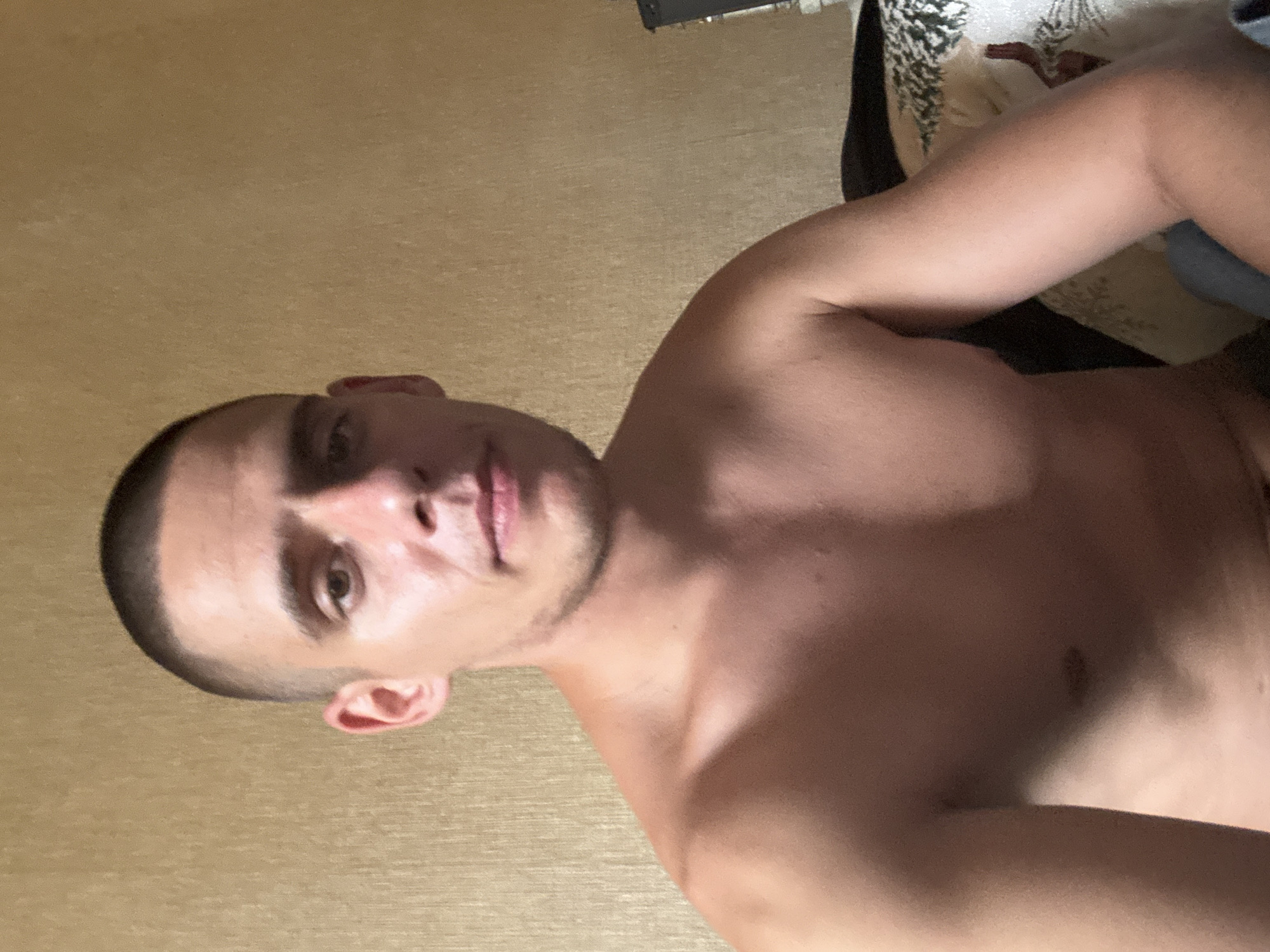 A young adult male with a shaved head and a fair complexion is looking directly at the camera, seated indoors. He is shirtless, revealing his chest and shoulders. The background is a textured, light brown wall. To the right of the man, a portion of a patterned cushion or blanket is visible, along with the edge of a dark object, possibly furniture. The overall lighting is dim, casting shadows on his face and body. The image appears to be a selfie taken in a residential setting. There is no visible indication of the specific location within Sofia, Bulgaria.