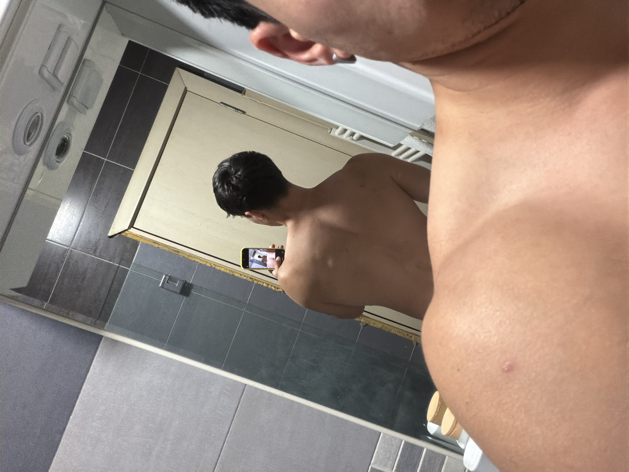 The media file captures a mirror selfie taken indoors, likely in a bathroom or changing area in Sofia, Bulgaria. The primary subject is a person, partially visible in a close-up on the right side of the frame, showing their right shoulder, upper back, and ear with an earring. A distinct tan line is visible across the shoulder and upper back. A small, reddish blemish is also present on the skin.
Reflected in the mirror, which occupies the left and central parts of the image, is the same person seen from the back. This person has short, dark hair and appears shirtless. They are holding a smartphone with a yellow case, using it to take the picture. The screen of the smartphone in the reflection shows an image that appears to be a person's torso or chest.
The setting features tiled walls in various shades of grey, dark grey, and lighter grey, with white grout lines. Part of a door frame is visible in the reflection, along with what seems to be a white bathroom fixture or part of a shower unit in the upper left of the reflection. Below the mirror, there's a surface (possibly a shelf or vanity) with a wooden tray and a few indistinct objects, potentially toiletries. The overall lighting suggests an indoor setting, likely with artificial light. No visible text is discernible in the image.