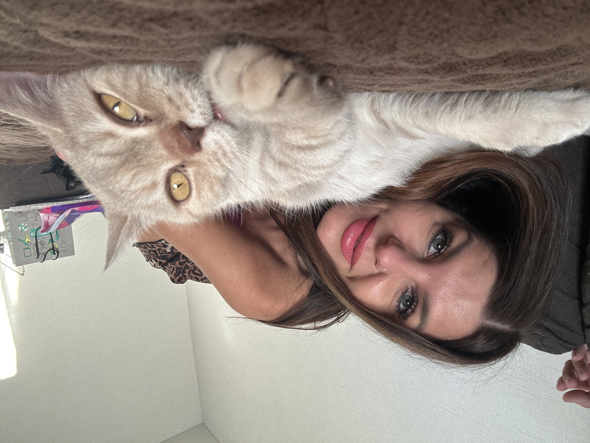 The image, taken in Chaikovsky, Russia, shows a woman and a cat lying closely together, suggesting a relaxed, intimate moment indoors. The perspective is a close-up, possibly a selfie, with both subjects oriented towards the camera.
The woman, with long, dark brown hair, has striking blue-green eyes and a subtle smile, enhanced by pink lipstick. She appears to be wearing a leopard-print top on her left shoulder and a darker garment on her right, with her right hand visible in the lower portion of the frame.
Beside her is a light fawn-colored cat with faint tabby markings. The cat's large, intense amber-yellow eyes with narrow pupils are fixed on the camera. One of its paws is visible near its head, appearing to be in a resting or playful position.
They are resting on a brown, textured surface, likely a blanket or sofa. The background features a bright white wall, subtly lit by natural light entering from the left. On the white wall, to the left of the woman's head, a partially visible, spiral-bound notebook or planner can be seen, adorned with colorful designs and the mirrored word "SCIENCE" printed vertically. The scene conveys a sense of calm and affection during the daytime.