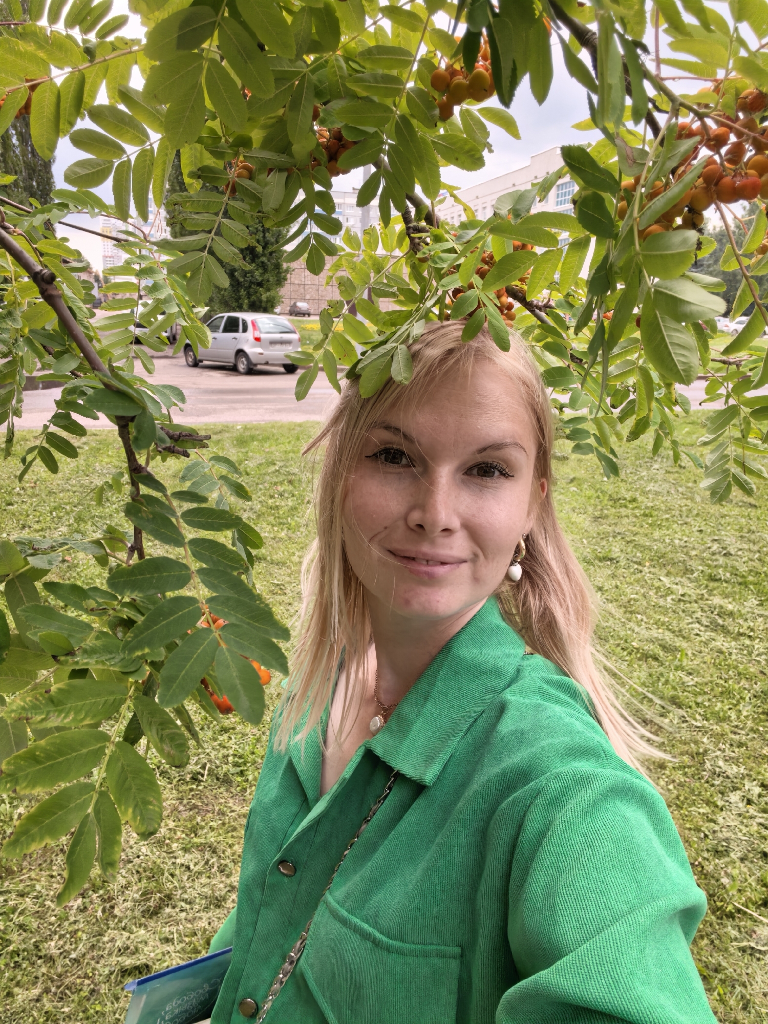 A young woman with blonde hair smiles at the camera, framed by the green leaves and orange berries of a mountain ash tree. She wears a green corduroy jacket and a necklace with a pearl pendant. She is outdoors, on a grassy area with a paved road and a silver car visible in the background. Buildings are also visible in the distance, suggesting an urban or suburban setting. The lighting suggests it is daytime. The woman's expression is happy and relaxed. No visible text is present in the image.