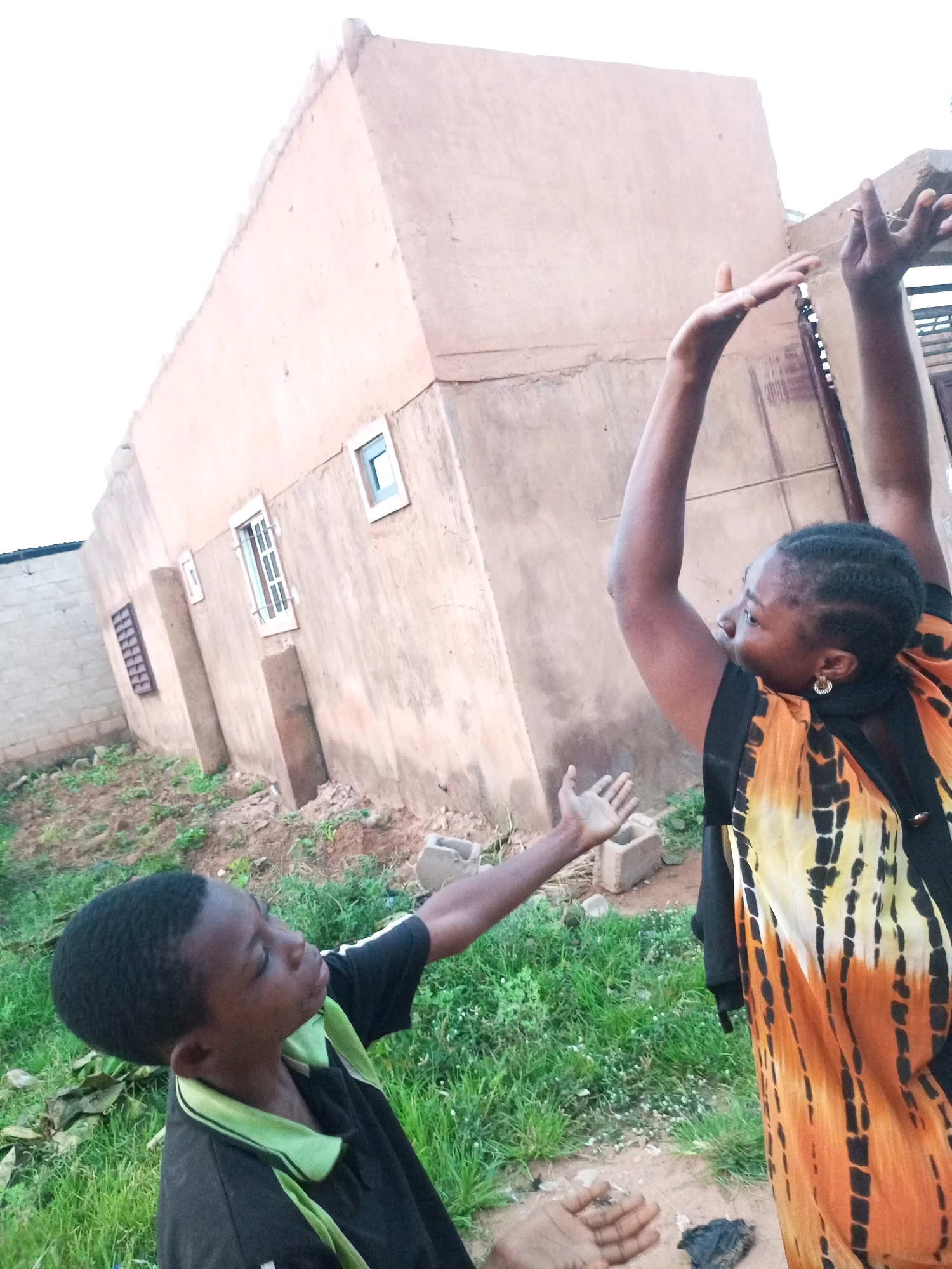 The image shows two individuals, a boy and a woman, in front of a building in Tempure, Nigeria. The building, possibly under construction, has a rough, unpainted facade and a few windows. The boy, wearing a black shirt with a green collar, is looking upwards and extending his left hand. The woman, in a brightly colored orange and black patterned garment, is also looking upwards with her arms raised. The ground around them is covered in dry earth and patches of grass. The sky above is overcast.