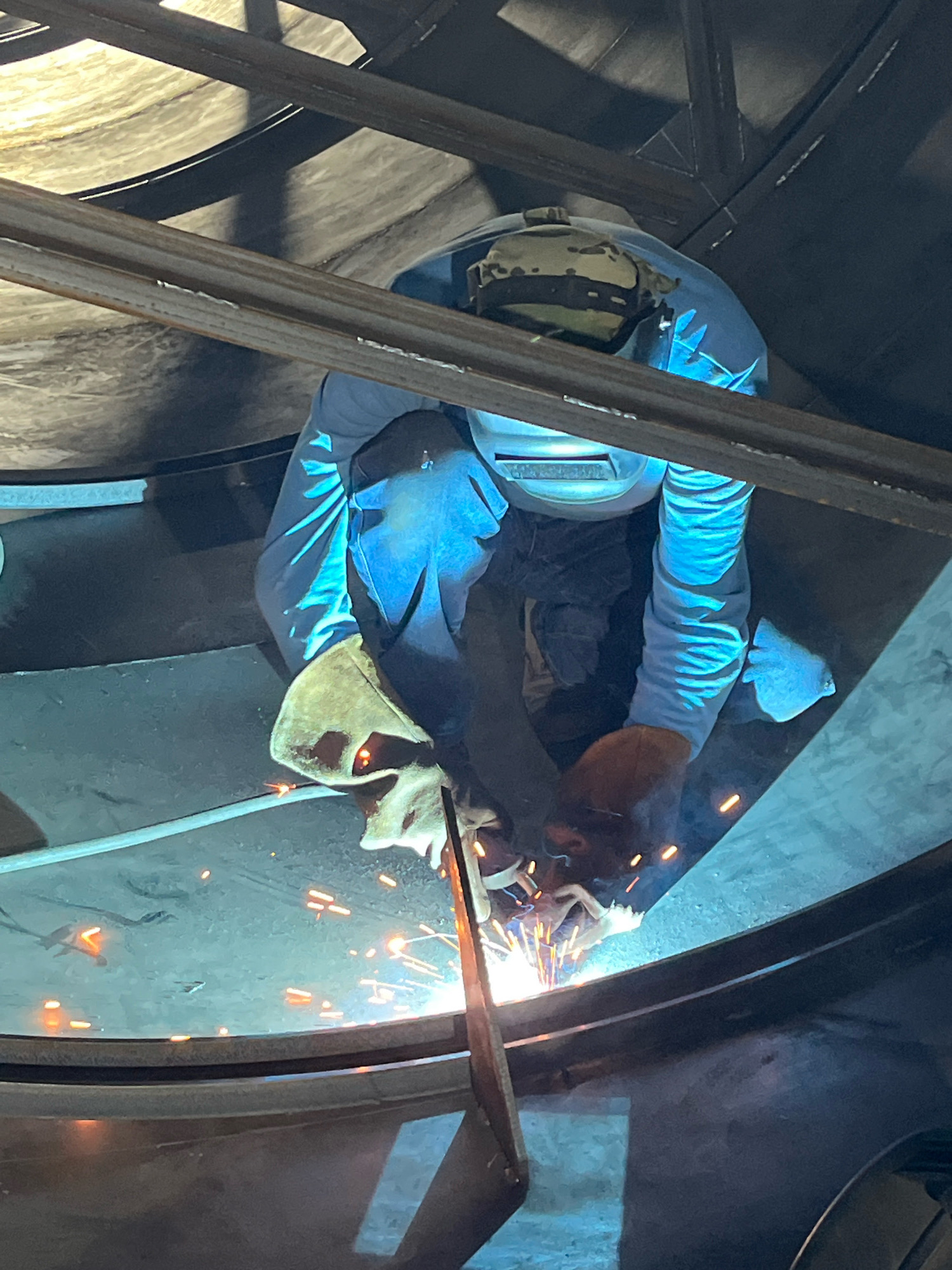 A welder, wearing a camouflage hard hat and protective gear, kneels inside a large cylindrical structure. The welder is actively welding a seam, indicated by the bright sparks and blue light emanating from the welding torch. Metal beams are visible overhead, suggesting an industrial or construction setting. The overall environment appears to be dark, with the welding light being the primary source of illumination. The image does not provide any specific details to determine the location in Taganrog City District, Russia.