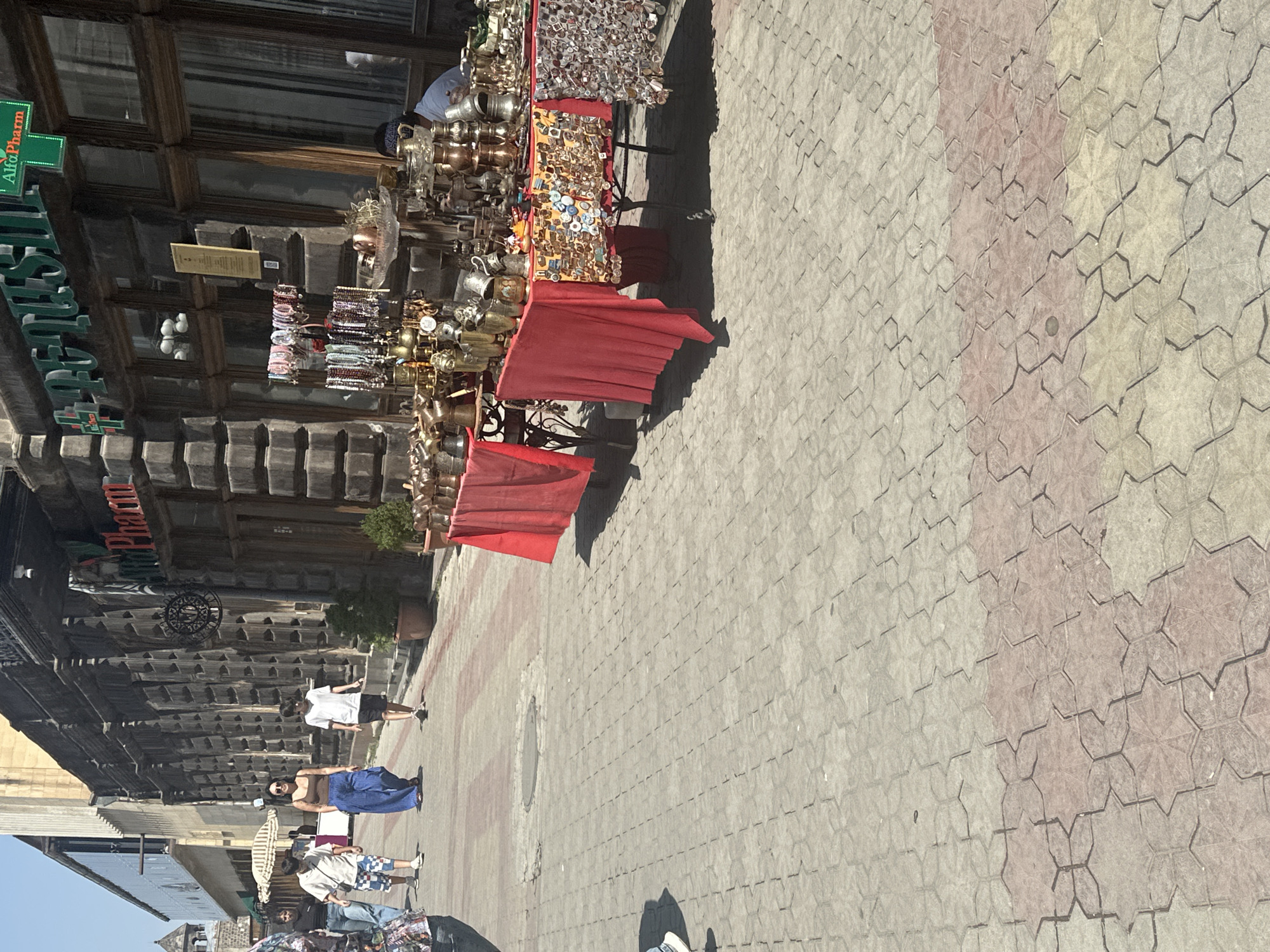 The image depicts a street scene in Gyumri, Armenia. On the right side of the image, a street vendor displays a collection of various items on tables covered with red fabric. These items appear to be souvenirs, including small metallic objects, possibly decorative pieces or trinkets, and some larger copper or brass vessels. Behind the vendor's stall, the facade of a building is visible, with architectural details and signage that include the words "Alfa Pharm" and "Pharcın". To the left of the vendor's stall, several people are walking along the street. Two young individuals, a boy and a girl, are walking side by side, with the boy wearing a light-colored shirt and patterned shorts, and the girl wearing a brown top and wide, blue pants. Further to the left, another person, likely a young boy, is walking away from the camera. The street is paved with interlocking concrete blocks, arranged in a patterned walkway. In the background, buildings with stone facades and windows can be seen, suggesting an urban environment. The overall impression is that of a sunny day in a commercial area where street vendors are present.