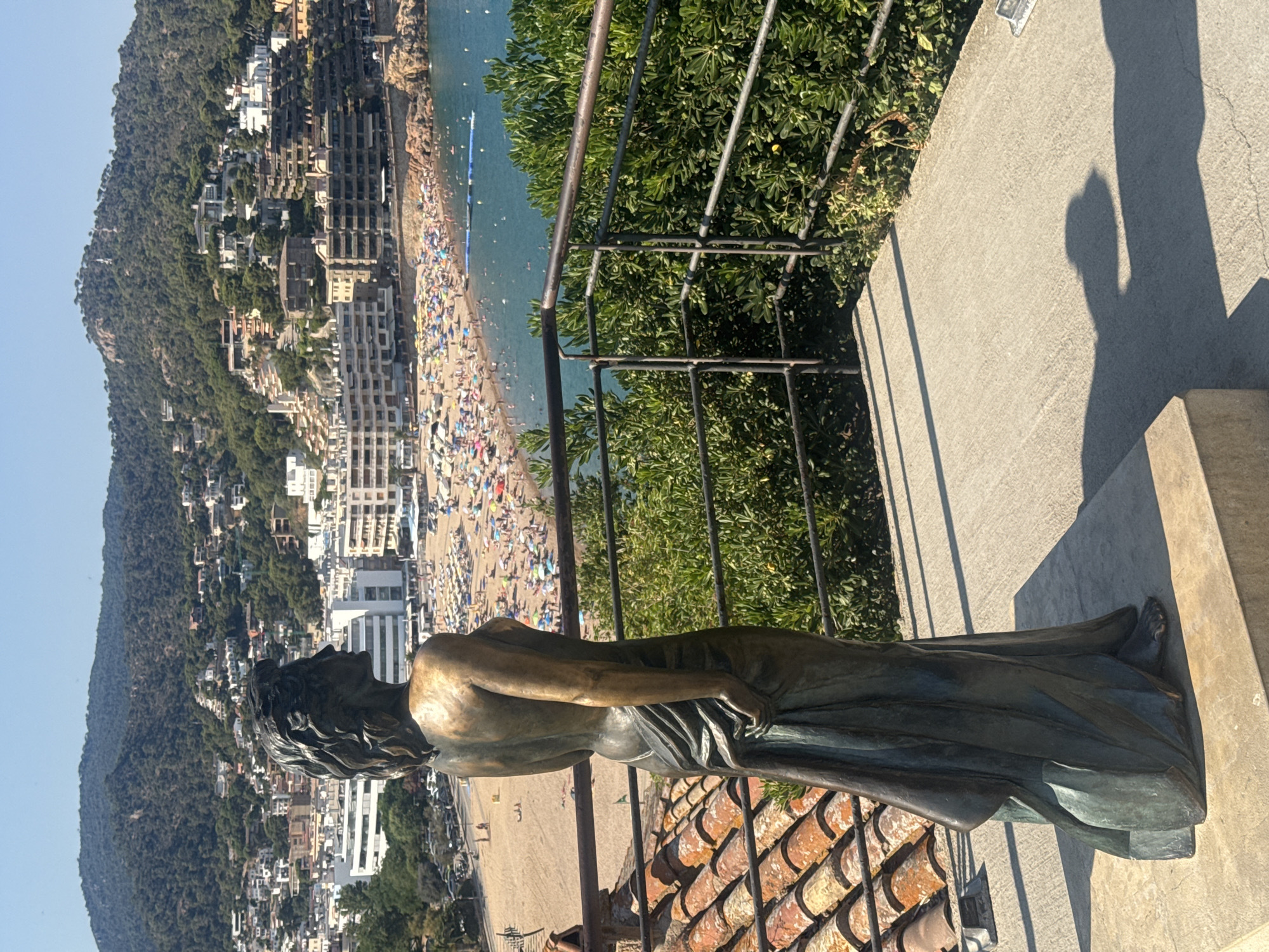 A bronze sculpture of a woman facing away from the viewer, her head turned to the left, is positioned on a concrete platform. She wears a draped gown. In the background, a bustling beach with numerous sunbathers and umbrellas lines a bay. Beyond the beach, a town with white buildings climbs a densely vegetated hillside, culminating in a mountain summit with a prominent radio tower. The foreground features a terra cotta tiled roof and a metal railing. The scene is illuminated by bright sunlight, casting distinct shadows. The location is identifiable as the viewpoint overlooking Platja Gran, Tossa de Mar, Spain, a popular coastal town on the Costa Brava.