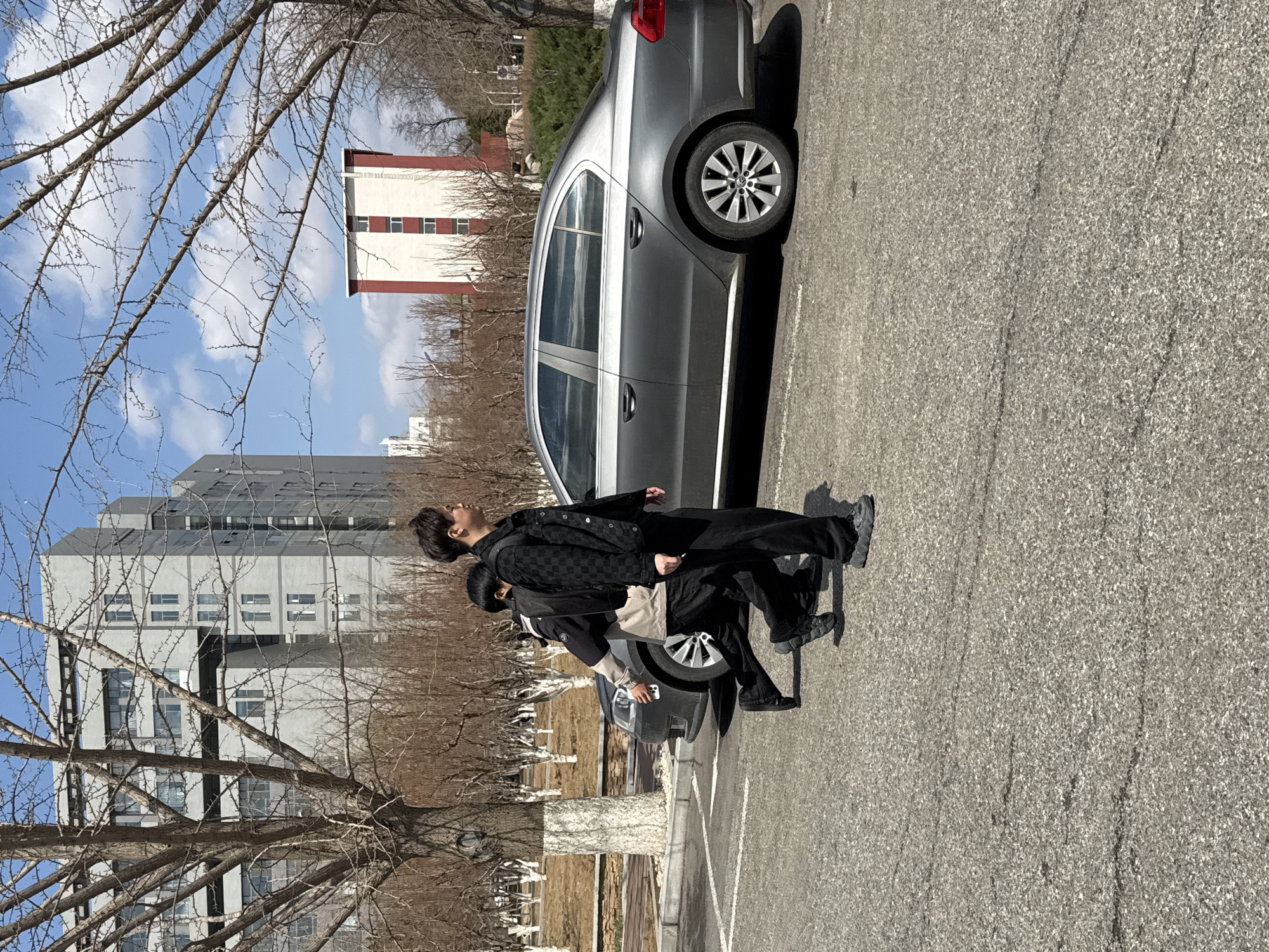 The image shows two people standing next to parked cars in Zhongtun, China. The taller person is wearing a black patterned jacket and black pants. The other person is wearing a black jacket with a tan bag. The background includes a tall building with many windows, as well as trees without leaves. The sky is blue. The area appears to be a parking lot or open space adjacent to buildings.