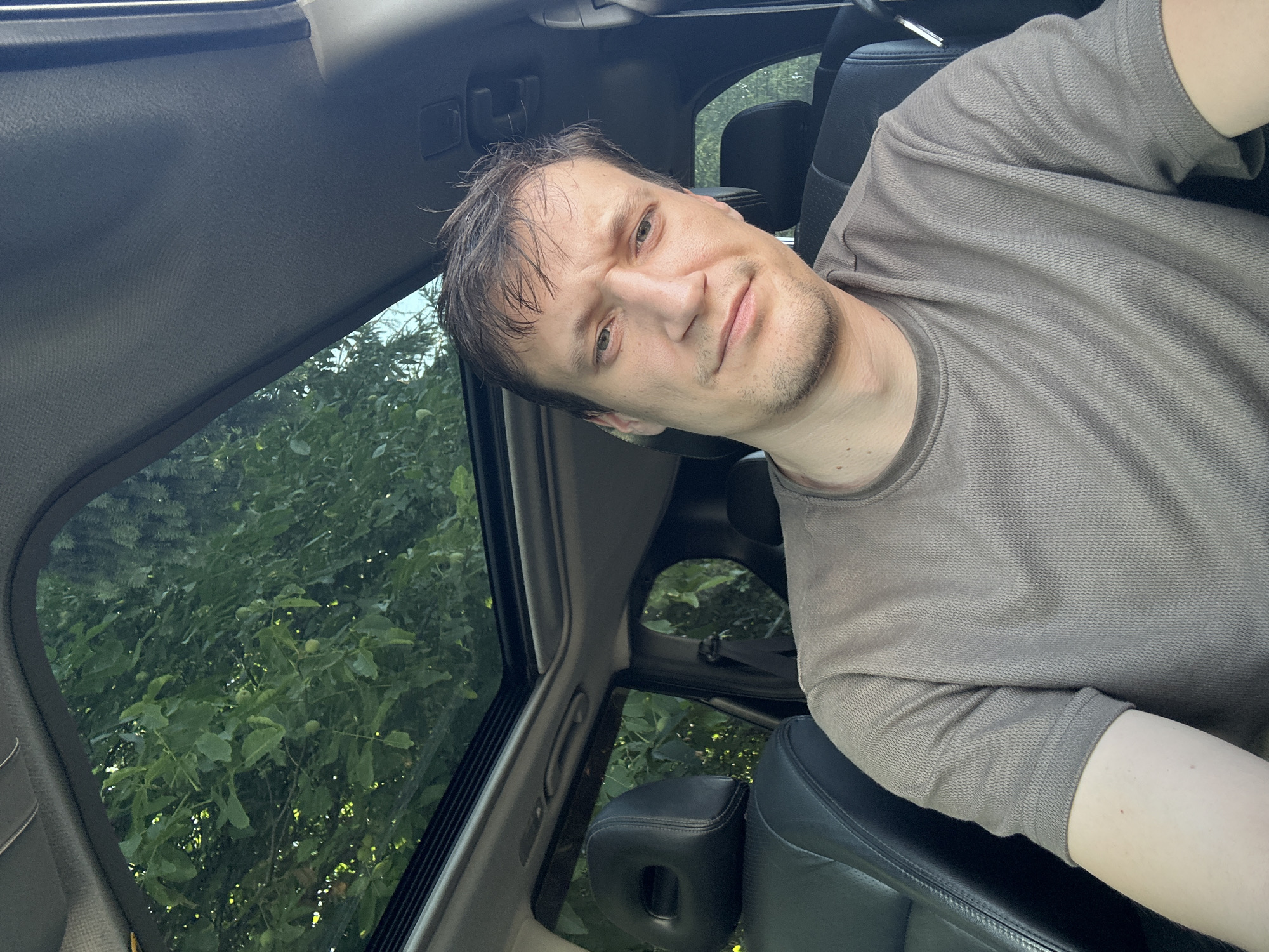 A selfie taken from the interior of a car shows a man with brown hair and a stubbled chin, wearing a taupe t-shirt. He is seated in the driver's seat, looking directly at the camera with a neutral expression. The car's sunroof is open, revealing a canopy of green trees and foliage above, suggesting an outdoor setting. The car's interior includes black leather seats and a part of the dashboard. The image appears to have been taken during daylight. The location is Shatava, Ukraine.