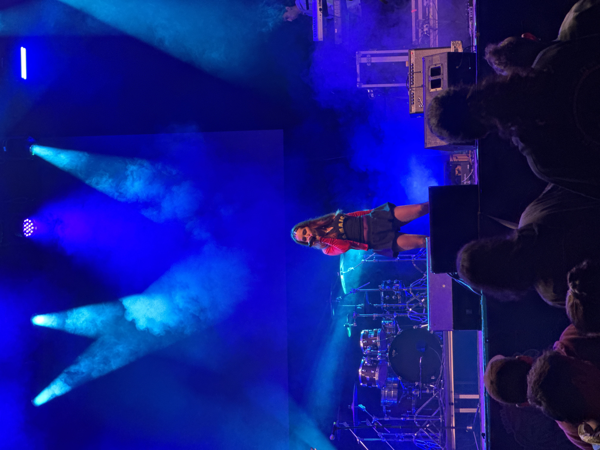 A performer with long brown hair sings into a microphone on a stage lit with blue lights. She wears a red jacket and black dress. Behind her is a drum set and other musical equipment. In the foreground, the heads and shoulders of audience members are visible. The location is Vilanova i la Geltrú, Spain, as implied by the source information.