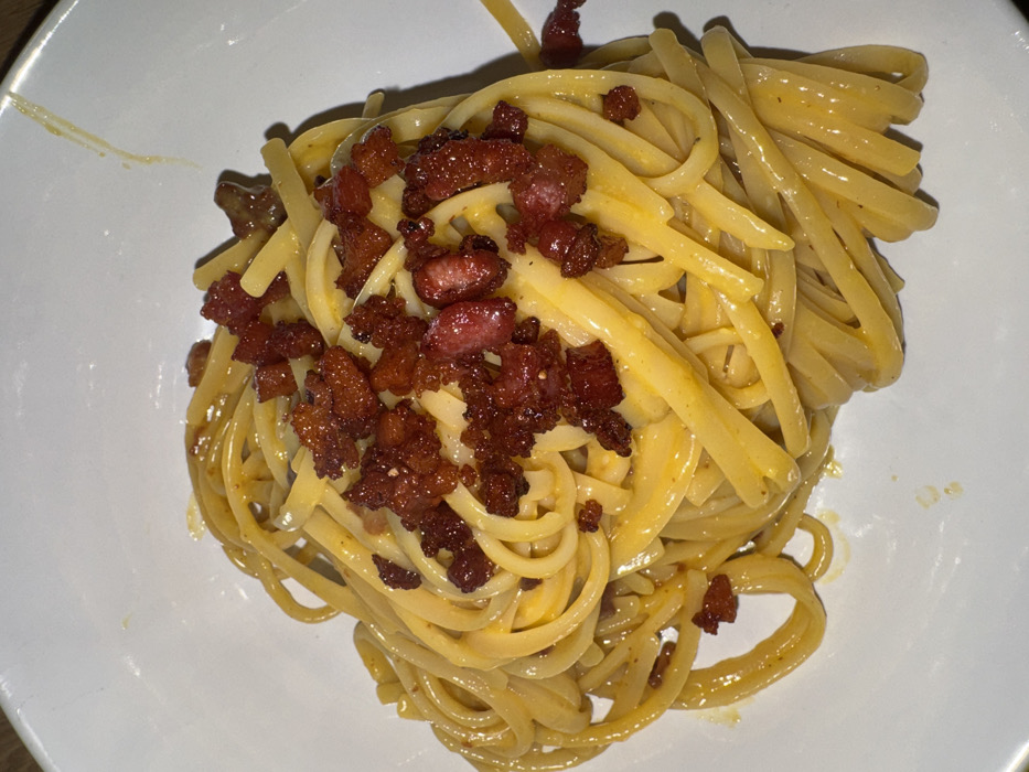 The image presents a close-up, overhead view of a pasta dish served in a white, shallow bowl or deep plate. The main subject is a substantial portion of long, flat pasta strands, consistent with linguine or fettuccine, uniformly coated in a viscous, pale yellow sauce. This sauce adheres to the pasta, giving it a glossy appearance. Scattered generously over the pasta are numerous small, irregular pieces of dark reddish-brown rendered meat, consistent with crispy pancetta or guanciale, some exhibiting lighter pinkish-red sections. A thin streak of the yellow sauce is visible on the upper right inner rim of the white dish. The lighting appears artificial, suggesting an indoor environment. No visual cues within the image confirm a specific location within Milan, Italy, or any other city.
