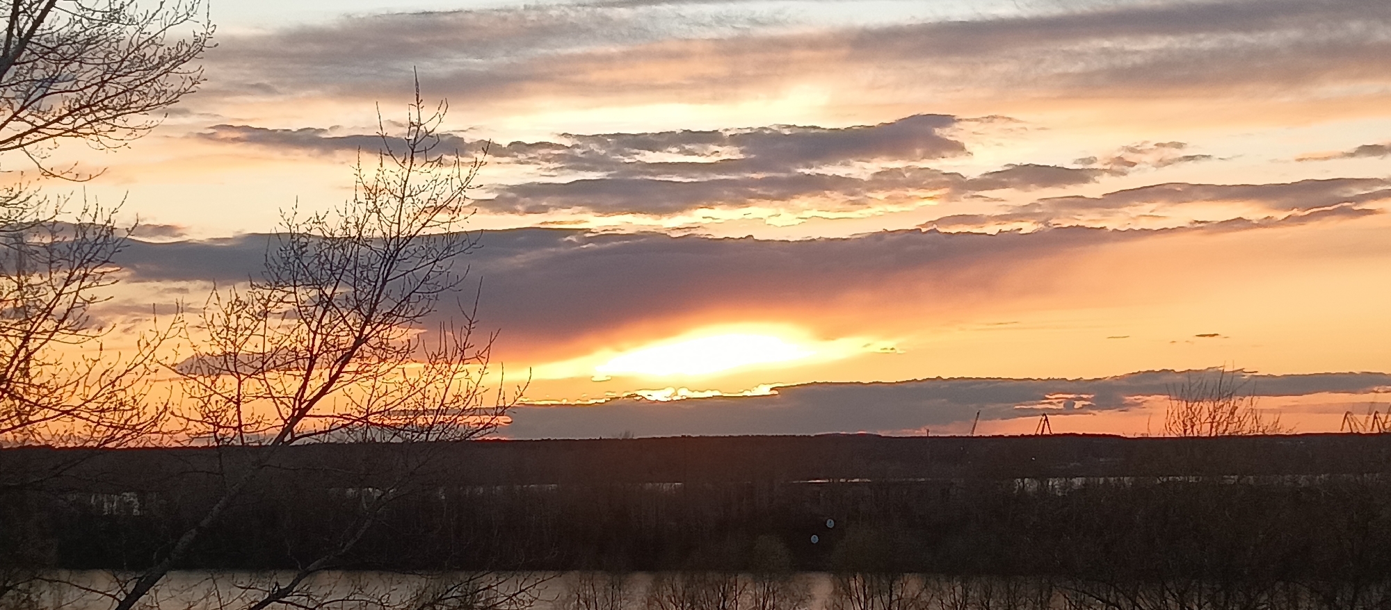 The media file displays a panoramic landscape captured during either sunrise or sunset. The sky, occupying the upper two-thirds of the image, is a prominent feature, filled with a dramatic array of clouds painted in warm hues of orange, yellow, and soft pink, contrasting with cooler gray and blue tones. The sun, a brilliant golden light, is partially obscured by a thick, dark band of clouds along the horizon, casting an intense glow that illuminates the edges of the surrounding cloud formations. Higher in the sky, thinner, stratified clouds are streaked across the vibrant expanse.
In the foreground, the intricate silhouettes of bare tree branches frame the view, indicating a late autumn or winter setting. Below the distant horizon, a dark, calm body of water, likely a river or lake, reflects the soft light from above. The horizon itself is a dark, undulating line of land or distant trees. Faint outlines of what appear to be industrial structures or cranes are visible on the distant right side of the horizon, hinting at a nearby urban or industrial presence.
The scene captures a tranquil moment of natural beauty, devoid of human activity or explicit subjects other than the landscape itself. The time of day is clearly dawn or dusk, marked by the low sun and rich sky colors. No visible text is present in the image.