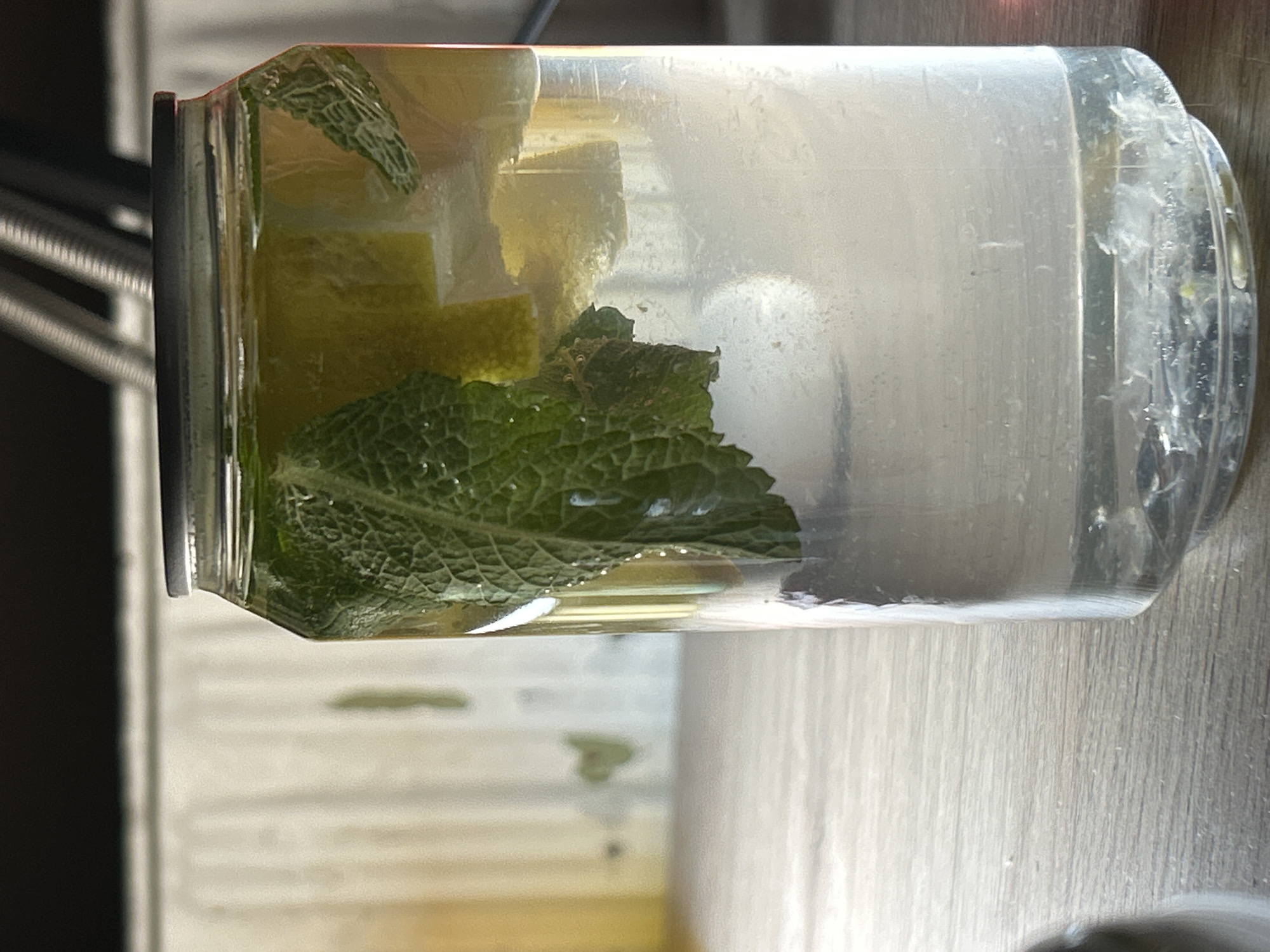 A clear glass jar containing water, lime wedges, and fresh mint leaves sits on a light-colored wooden surface. The jar is upright and oriented with the lid to the left of the frame. The lighting is diffused. The scene appears to be indoors, with a portion of a black object (possibly a table or chair) visible in the background on the left. The precise location in Sofia, Bulgaria is not discernible from the visual information.