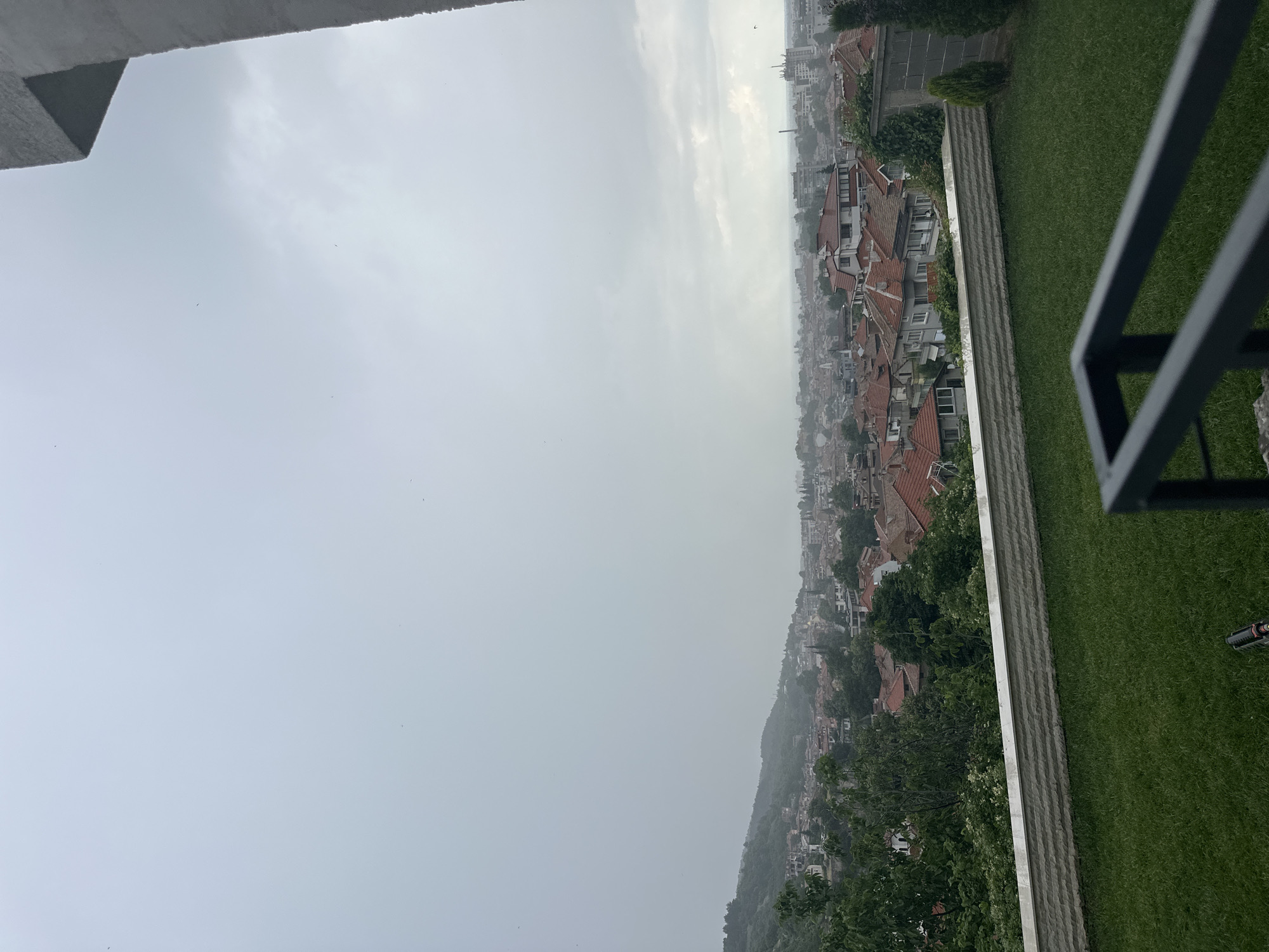 The media file is an image taken from a high vantage point, looking out over a city and a cloudy sky. The camera is oriented such that the scene is vertical.
In the foreground, there is a glimpse of a grey railing and a lush green lawn. Beyond the lawn, a stone retaining wall separates it from a densely populated city area. The city features many buildings with reddish-brown tiled roofs, interspersed with trees. The buildings appear to be of varying heights, suggesting a historical and residential urban landscape. The city sprawls across a hillside, with buildings extending up and down the slopes. In the far distance, the cityscape fades into a hazy, overcast horizon.
The sky is completely overcast with grey clouds, indicating a cloudy or possibly rainy day. The weather appears to be dull and muted, with a lack of direct sunlight. The overall mood conveyed by the image is somewhat somber or tranquil due to the weather and the expansive, somewhat distant view of the city.
No people are visible in the image. There are no discernible objects apart from the architectural elements and the natural landscape. The image is likely taken during the daytime, given the illumination, but the overcast conditions prevent a precise determination of the time of day. The location cues point to Asenovgrad, Bulgaria, as indicated by the context provided. There is no visible text in the image.