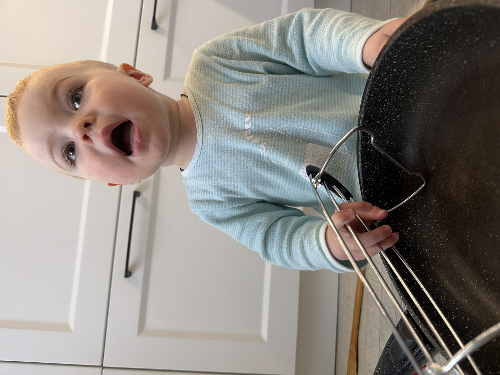 A young child with blond hair and blue-grey eyes is shown indoors, appearing to be in a kitchen. The child, wearing a long-sleeved, light blue and white striped shirt, has their mouth open in a possible expression of surprise or excitement. The child is holding a black frying pan with a silver handle. The background features white cabinets with black hardware. The exact location in Votkinsk, Russia, is not discernible from the image.