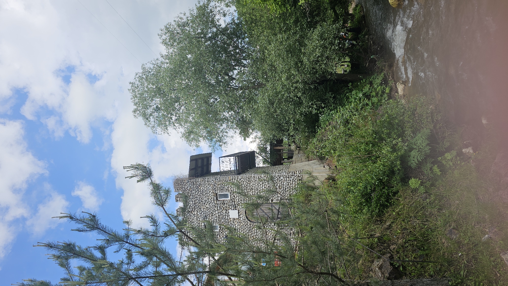 The image depicts a stone structure resembling a small tower or fortification, constructed with rounded stones. It has a balcony with a metal railing. Adjacent to the structure, a large, leafy tree dominates the right side of the frame. A pine tree is visible in the foreground on the left, partially obscuring the stone building. A body of water, likely a river or stream, flows in the lower right portion of the image, with white water indicating a current. Lush green vegetation and trees surround the scene, suggesting a natural or semi-natural environment. The sky above is blue with scattered white clouds. There are no people visible in the image. The overall impression is of a rustic or historical setting integrated with nature.