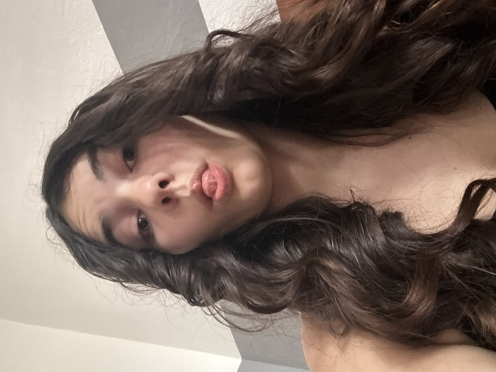 A selfie of a woman with long, dark wavy hair and light skin. The background consists of a wall and a ceiling corner painted in white and light gray. The woman is wearing an earbud in her left ear. Her lips are slightly parted, and her tongue is visible. The location is not discernible from this image; it cannot be determined if the location is in Plovdiv, Bulgaria.