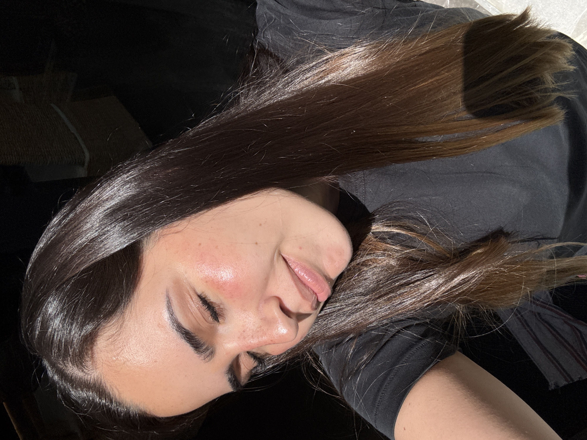 A young woman is lying down, with her eyes closed and a slight smile on her face. Her head is resting on her arm, and her long, dark brown hair is spread out around her. She is wearing a black t-shirt. The lighting is uneven, with some areas in shadow and others brightly lit. The background is mostly dark and obscured, but there is a hint of a light-colored object in the upper left corner. The setting is unknown, and cannot be identified.