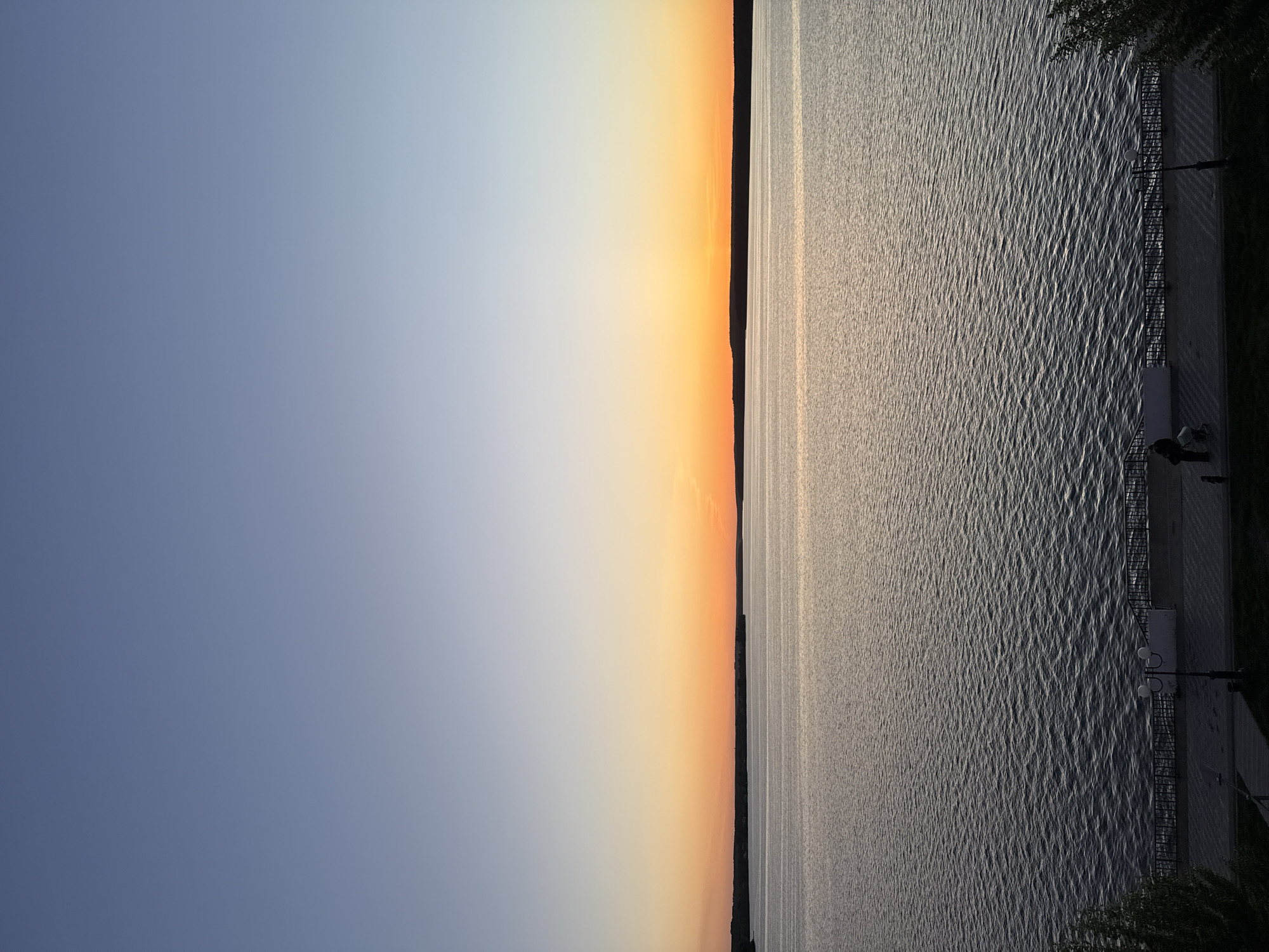 The image captures a view of the Votkinsk Reservoir in the city of Votkinsk, Russia, during a time near sunset. The sky transitions from a light blue on the left to orange hues near the horizon. The reservoir occupies the right side of the image. The water reflects the light and sky creating a textured surface. A shoreline with what looks like a paved walkway or embankment is visible along the right edge of the reservoir. Some street lights are situated along this path. A few silhouetted trees border the path. Two people are near the street lights, though their activities are indistinguishable.