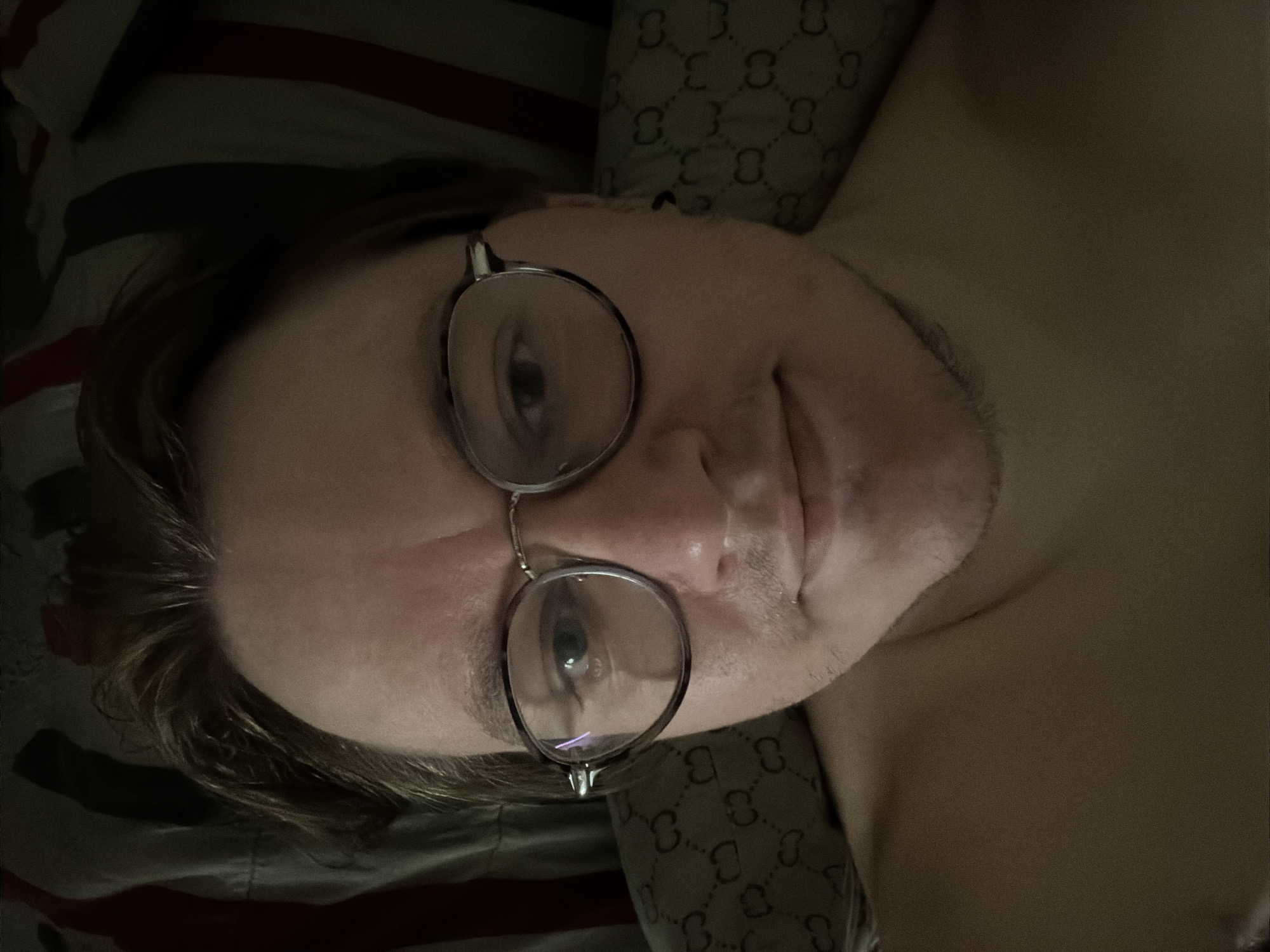 A close-up, overhead, natural light selfie shows a person wearing glasses and lying down. The person has fair skin, light brown hair, and appears to be a male. He is looking directly at the camera with a slight smile. The background consists of pillows with a striped pattern and a pillow with a repeating 'GG' logo. The person is shirtless.