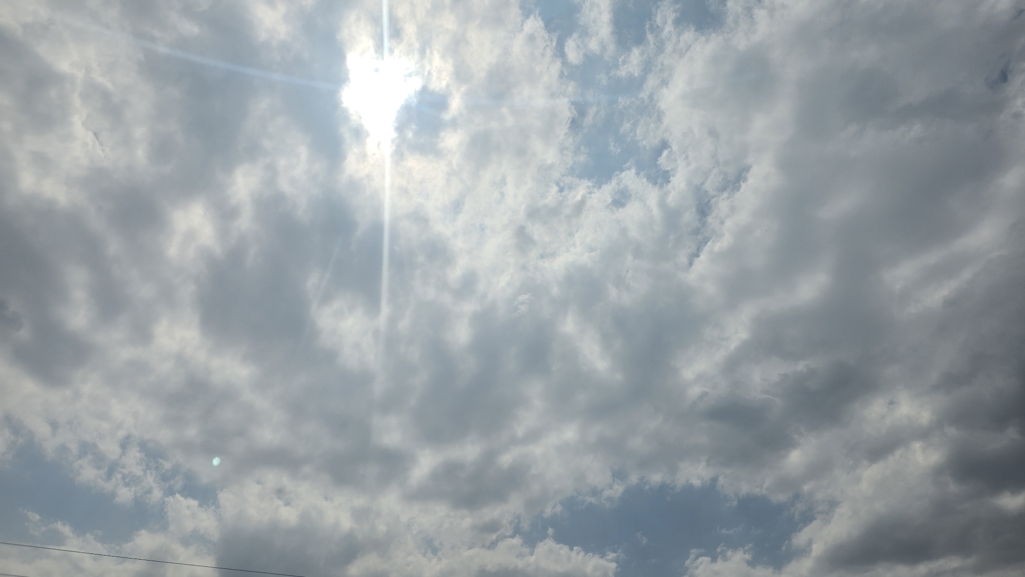 A wide shot captures a cloudy sky with the sun peeking through, creating a lens flare effect. The sun's rays are visible, streaking across the sky. The clouds are varied in density, with some appearing light and wispy, while others are darker and more substantial. The overall impression is of a partly cloudy day, possibly with some atmospheric haze, contributing to the diffused sunlight. The image does not depict any identifiable landmarks of Taldyk, Kyrgyzstan, or any people or specific objects within the frame.