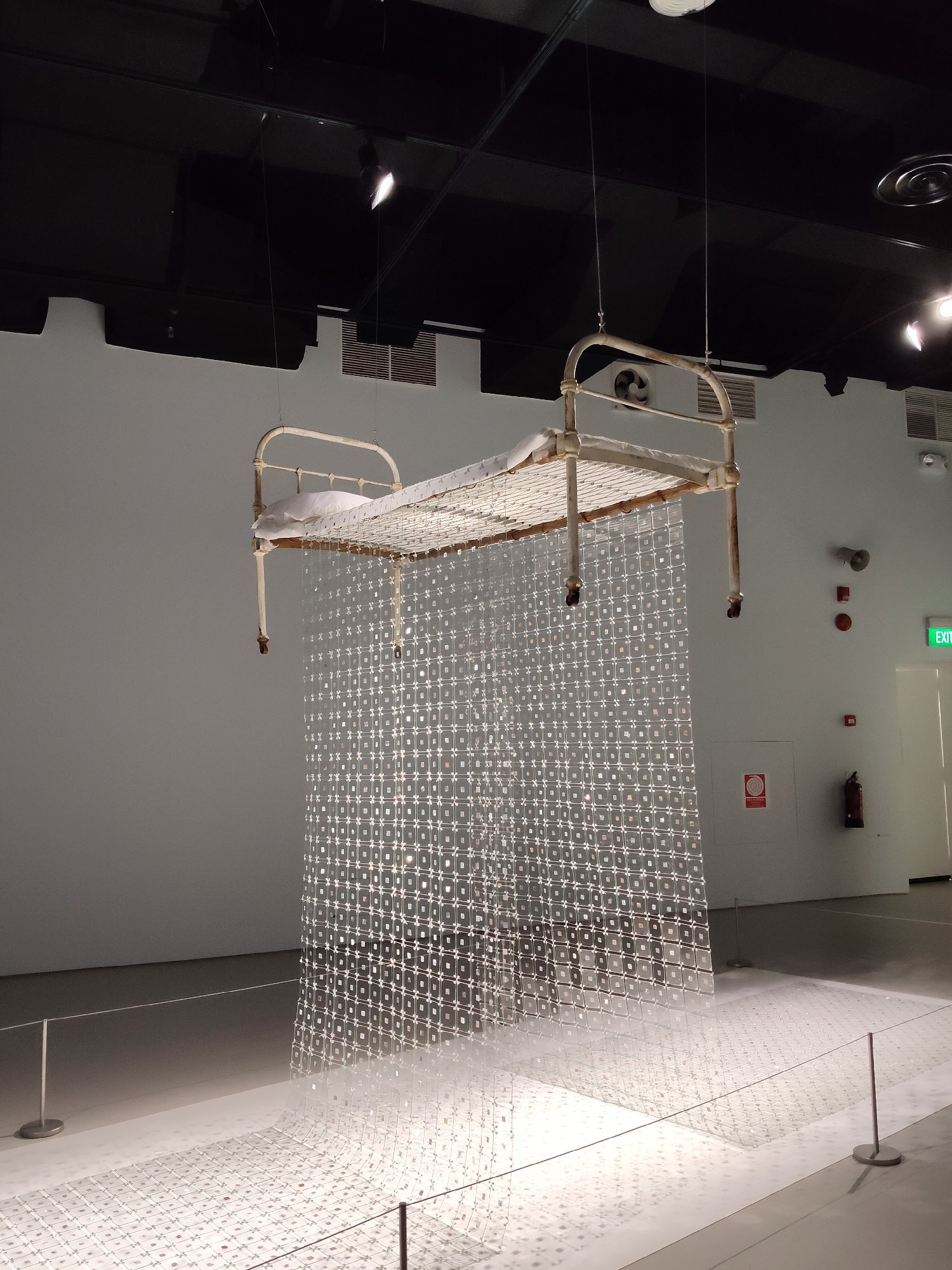 This is a photograph of an art installation in a museum in Singapore. The artwork consists of an antique-looking metal bed frame suspended from the ceiling by wires. The bed has a white mattress and a white pillow. Hanging from the bed frame is a curtain made of many small, clear square elements that create a shimmering, reflective effect. This curtain extends down to the floor, where it appears to spread out and cover a portion of the floor. The installation is in a large, well-lit room with a white wall and a dark ceiling. There are ventilation grilles and lighting fixtures visible on the ceiling. On the wall to the right, there is a green "EXIT" sign, a fire extinguisher, and some circular warning signs. A rope barrier is set up in front of the installation. The floor is white and reflects the light from the artwork. There are no people visible in the image. The overall impression is one of an ethereal, almost dreamlike display.