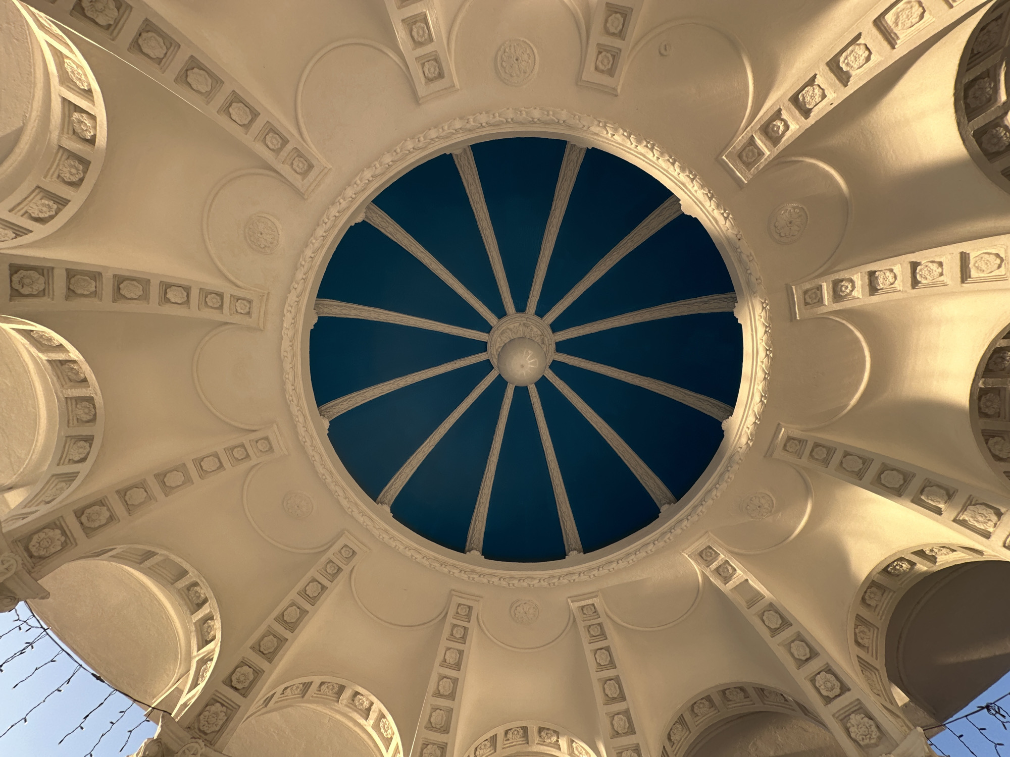 A worm's eye view reveals the intricate architecture of a structure with a blue dome, likely located in Sochi, Russia. The dome is circular with ribs radiating from a central point and light fixture. The surrounding structure is made of white or light-colored material, likely stone or plaster, and features repeated patterns of squares and circles with floral or ornate designs. Arches and columns are visible.