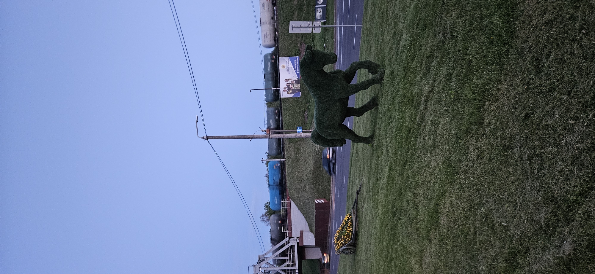 In Vitsebsk, Belarus, a decorative topiary sculpture of a horse stands on a grassy median beside a road. Behind the sculpture, a train of tank cars travels on elevated tracks. To the left of the horse sculpture, a wooden cart filled with yellow flowers is visible. A street lamp and power lines are present in the background, as well as a billboard with images of people and text in Cyrillic. Cars are seen driving on the road. The sky is clear and blue, indicating daytime.