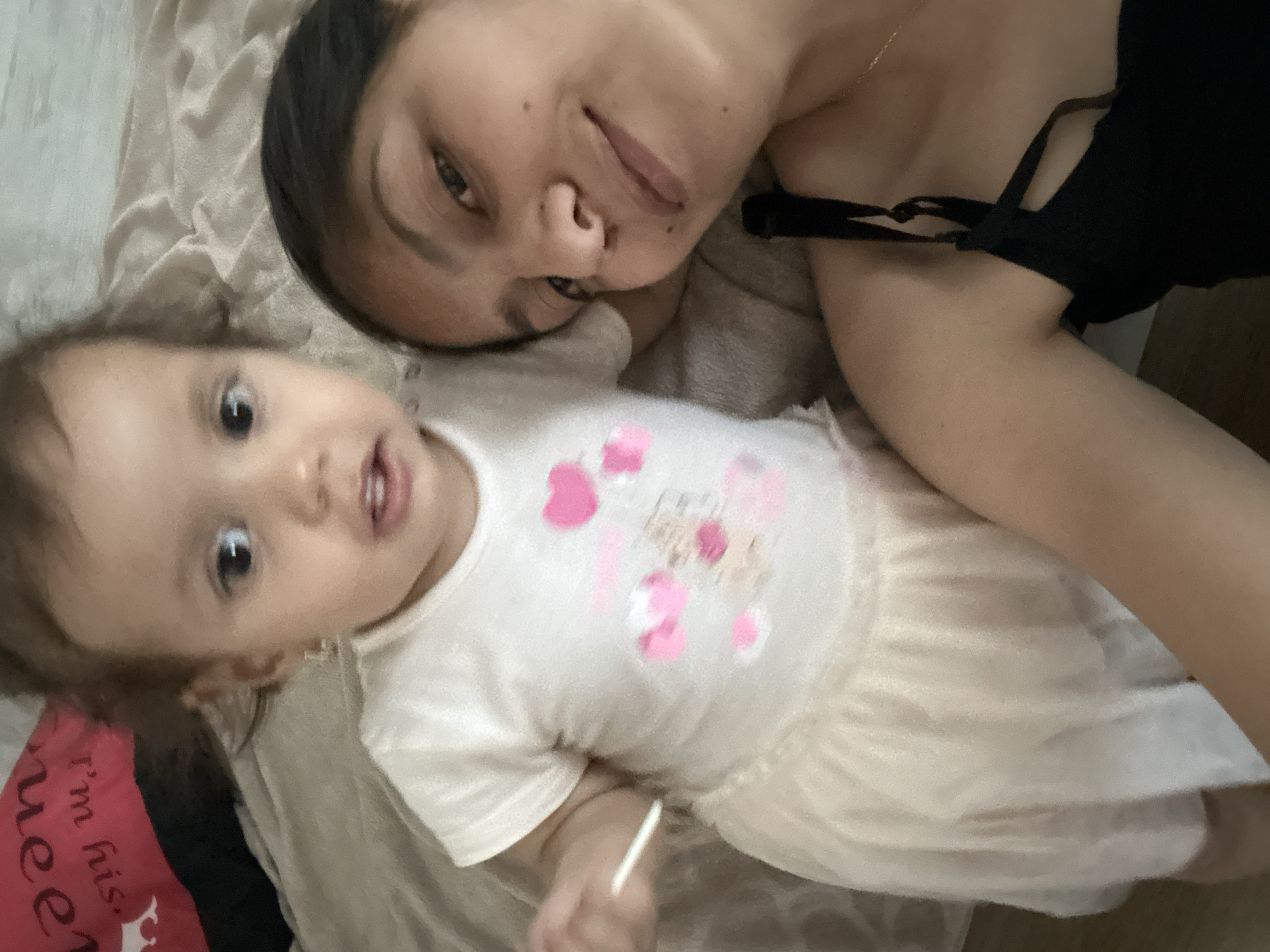 The image shows two individuals, an adult female and a young child, lying on a bed. The adult female, in the foreground and on the right side of the image, is looking towards the camera with a slight smile. She is wearing a black top with thin straps. The child is positioned behind and to the left of the adult, looking towards the camera as well. The child is wearing a white t-shirt with pink heart and cartoon graphics, and a light-colored skirt. A pink pillow with text is visible behind the child. The background appears to be a light-colored bedspread. The image does not contain any information that would allow for the determination of its location in the city of Parvomay, Bulgaria.