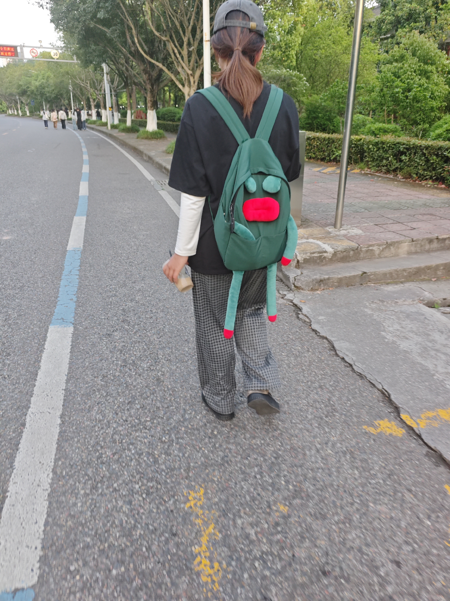 A person is walking on the side of a road, as seen from the rear view. The individual, likely a woman, has a ponytail, wears a gray baseball cap, black shirt over a long-sleeved white shirt, and checkered pants. She carries a green backpack with a cartoonish face. The person is walking along a road marked with blue and white lines and holding a cup. The location appears to be an urban street in Chongqing High-tech Zone, China, with trees lining the roadside and other people visible in the distance.