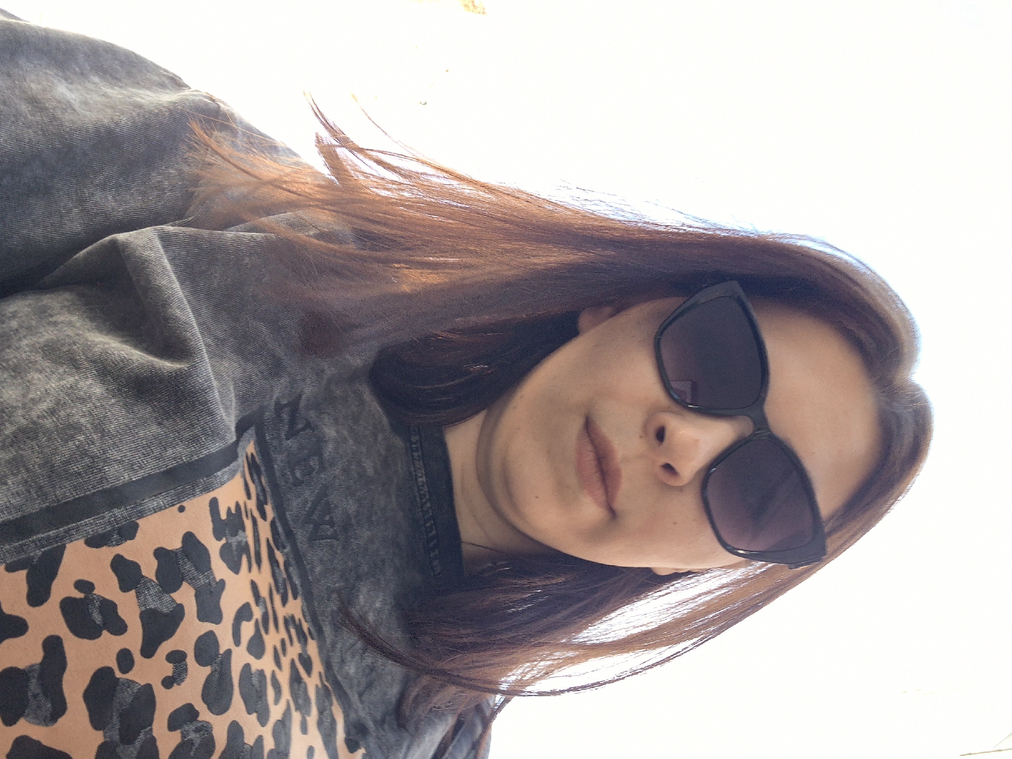 The media file presents a close-up portrait of a person, likely a woman, taken outdoors in Chaikovsky, Russia. The image is captured from a low angle, looking upwards, and is rotated almost 90 degrees counter-clockwise, with the subject's head tilted significantly.
The subject has long, dark brown hair, appearing with reddish highlights due to the bright lighting. She is wearing stylish dark sunglasses with angular black frames, obscuring her eyes. Her facial expression is neutral and relaxed, with her lips slightly parted.
She is dressed in a dark grey garment that exhibits a textured, possibly ribbed or corduroy, pattern. Below this, a prominent section of a tan and black leopard print garment is visible. A thin black band separates the grey fabric from the animal print. On the right side of the grey fabric, near the leopard print, there is some faint, distressed white lettering, but it is not clearly legible.
The background is almost entirely overexposed and white, indicating a very bright outdoor setting, likely against the sky or a brightly lit wall, signifying daytime. The overall impression is a casual, sunlit portrait or selfie. No specific architectural or landscape features of Chaikovsky are discernible in the background.
