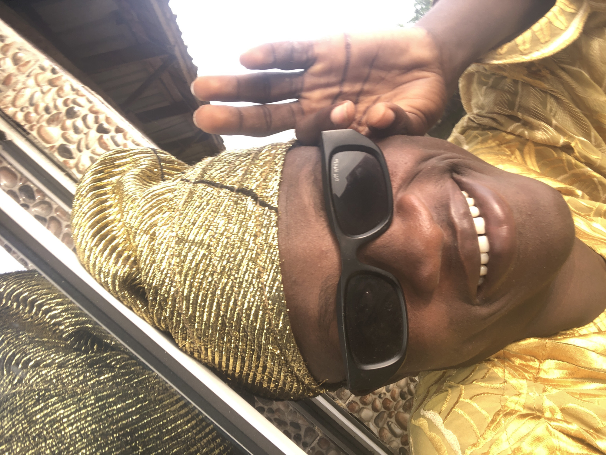 A close-up selfie captures a dark-skinned person wearing a gold, textured cap and a matching gold-embroidered outfit. The person has on black sunglasses with "off white" written on the left lens. They are smiling widely, showing teeth, and holding up their right hand, fingers spread, to their right temple. The background is blurred but shows a textured wall, possibly stone, and an overcast sky. The setting appears to be outdoors, with the bright sky suggesting daytime. The overall impression is of a candid, celebratory, or fashionable portrait. This image is not explicitly tied to Ado, Nigeria, but the attire and setting could be consistent with cultural expressions found there.