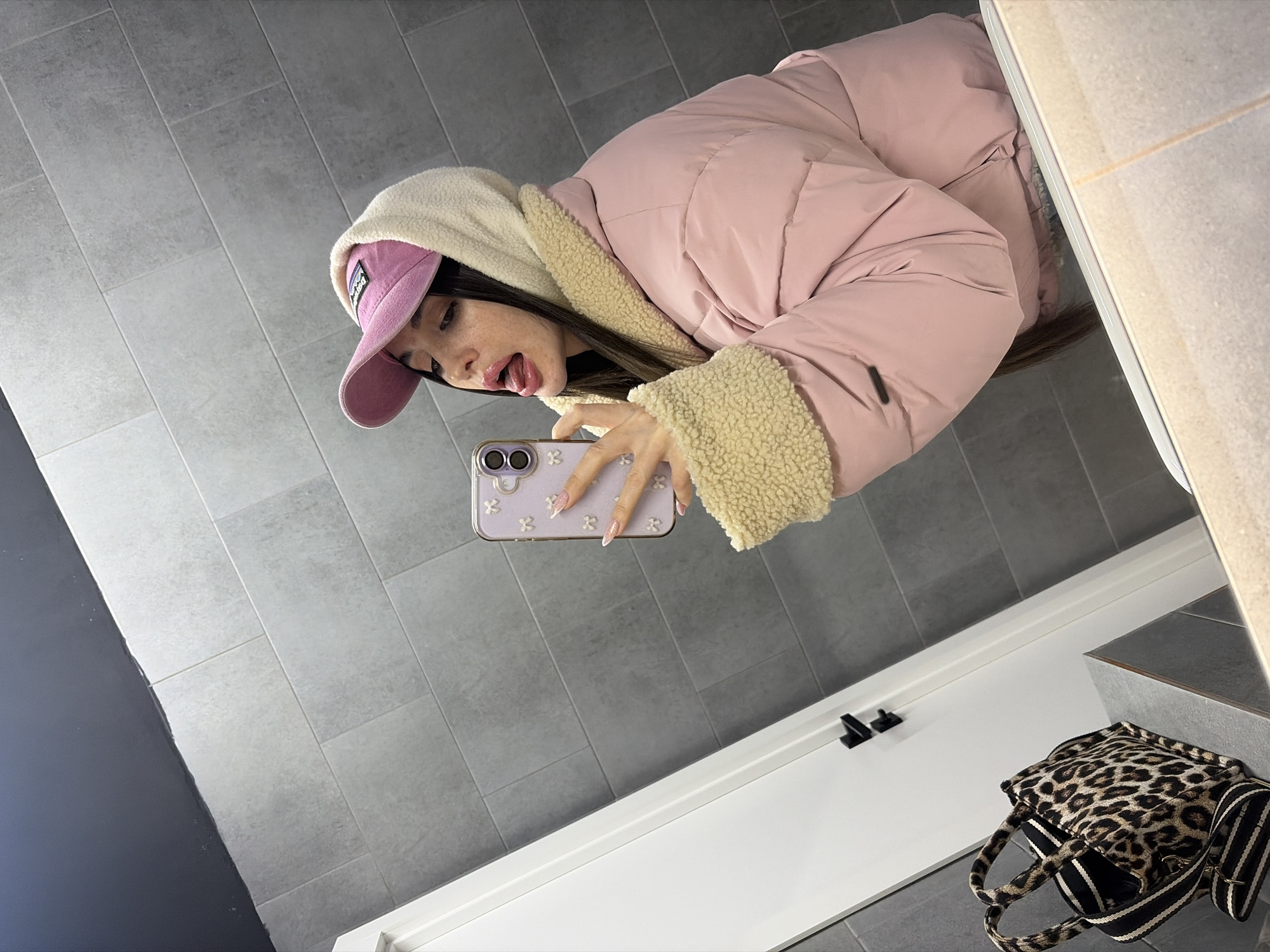 A young woman in a pink baseball cap and a pink puffer jacket with a beige sherpa lining is taking a selfie in what appears to be a bathroom. The walls are tiled in gray. A white door with black hardware is visible in the background. A leopard-print tote bag is on the floor near the wall. The location cannot be verified as Saint Petersburg, Russia, with the given information.