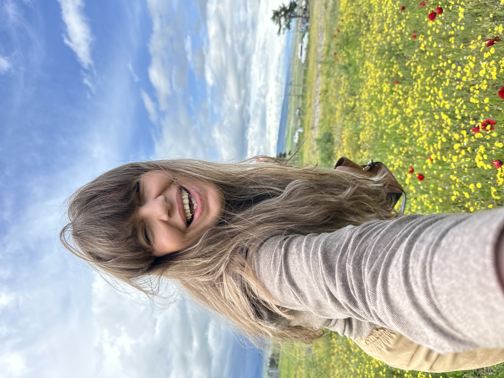 A smiling woman with long brown hair takes a selfie in a field of yellow and red flowers. The background consists of a blue sky with white clouds, a distant landscape with green fields, trees, and a few buildings. The photo appears to be taken at an angle, with the woman's head tilted. The exact location in Marneuli, Georgia, cannot be determined without more specific visual information.