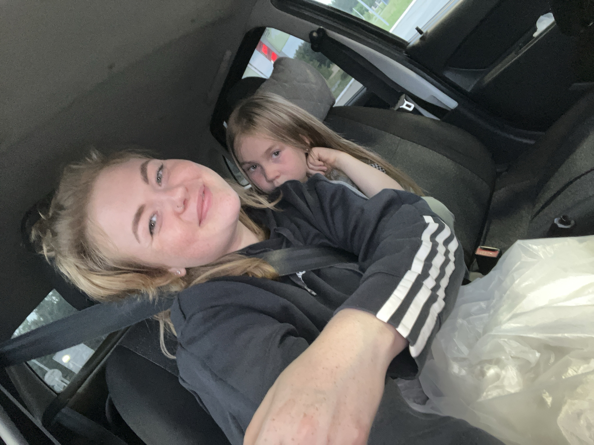 This is a close-up, selfie-style, medium shot image taken from the interior of a car. Two individuals, an adult female and a young girl, are the primary subjects. The adult female is in the foreground, seated in the driver's or front passenger seat, with her body angled towards the camera. She has blonde hair pulled up in a bun, is wearing a dark hooded sweatshirt with white stripes on the sleeves, and has a nose ring. She is smiling and looking at the camera, with her right arm extended towards the camera. She is wearing a seatbelt.
Behind and to the right of the adult female is a young girl, also seated in the car, likely in the back seat. She has long blonde hair and is looking towards the left side of the frame, resting her head on her hand. She is wearing a grey shirt and appears to be looking at something outside the car. The car's interior is visible, including the seats and a seatbelt buckle. A white plastic bag is visible in the lower left quadrant of the image, partially obscuring the adult female's lap. The background outside the car appears to be a somewhat overcast day with some greenery visible. There are no discernible landmarks or details to indicate the specific location of Chaikovsky, Russia, within this image.