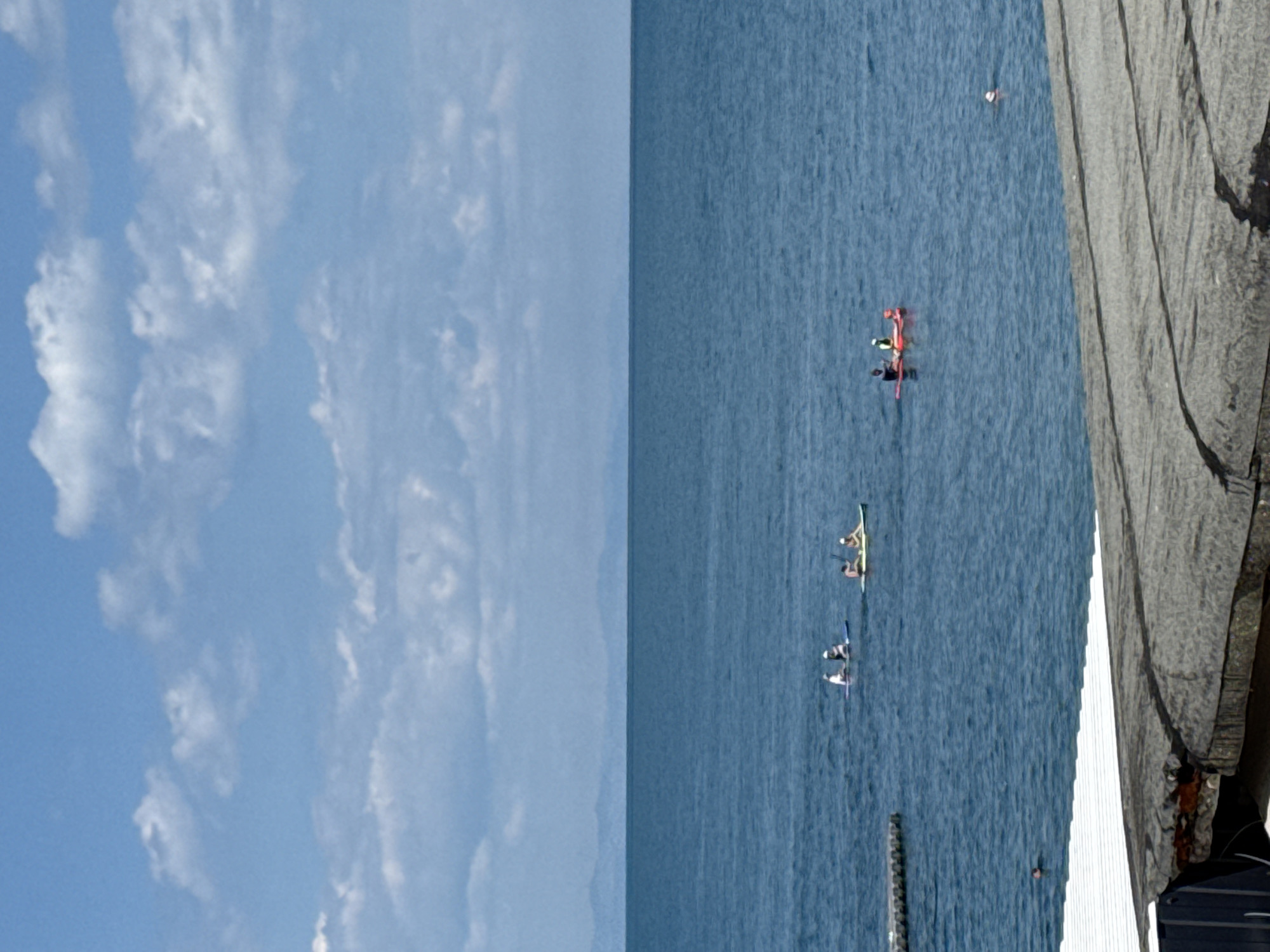 The image depicts a serene coastal scene with a vast expanse of deep blue ocean under a partly cloudy sky. In the middle ground, several individuals are engaged in watersports. One group of three is on paddleboards, while another individual is on a kayak. Further out, a single person can be seen swimming. In the foreground, a section of a weathered, flat roof or structure partially obscures the view of the water. The horizon is marked by a distant, hazy shoreline.