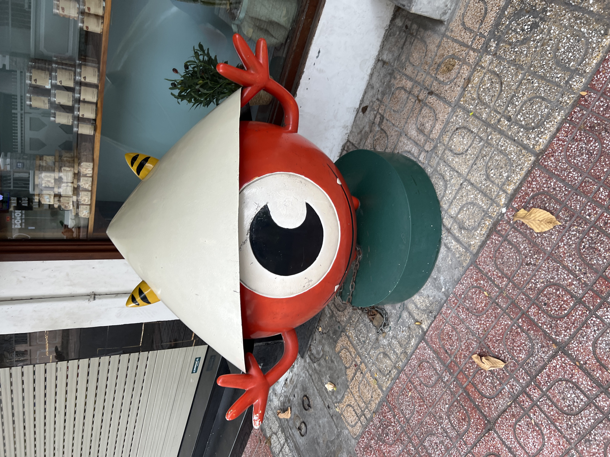 This is a medium shot of a whimsical, red, spherical statue with a large black and white eye, wearing a conical Vietnamese hat. The statue has two red, stick-like arms with four fingers each, raised slightly. Two yellow, black-striped horns protrude from under the hat. The statue is positioned on a green cylindrical base, which is secured to the ground by a metal chain and padlock. The statue stands on a patterned tiled sidewalk in front of a building with a closed, corrugated metal roll-up door and a shop window displaying jars of goods. The scene is located on a sidewalk in Nha Trang, Vietnam, with fallen leaves scattered on the pavement. The overall tone is vibrant and slightly surreal.