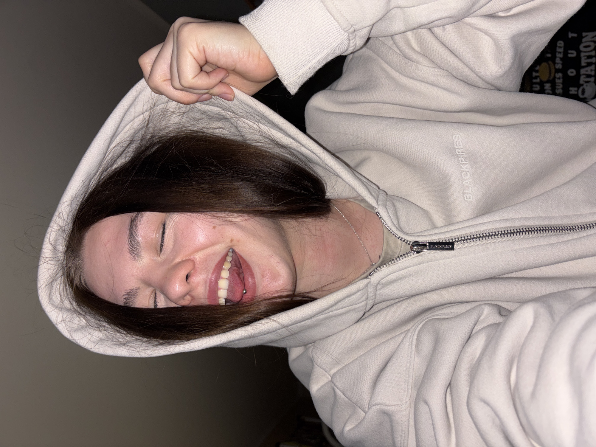 A young woman with brown hair is smiling broadly, her eyes nearly closed. She is wearing a cream-colored hoodie with the word "BLACKPIRES" printed on the left side. The hoodie is partially zipped and the hood is pulled up, framing her face. A silver chain is visible around her neck. She has a tongue piercing. The background is dark and indistinct, but a portion of a black garment with text that reads "STATION" is visible. The city of Perm, Russia, is not discernible in the image.