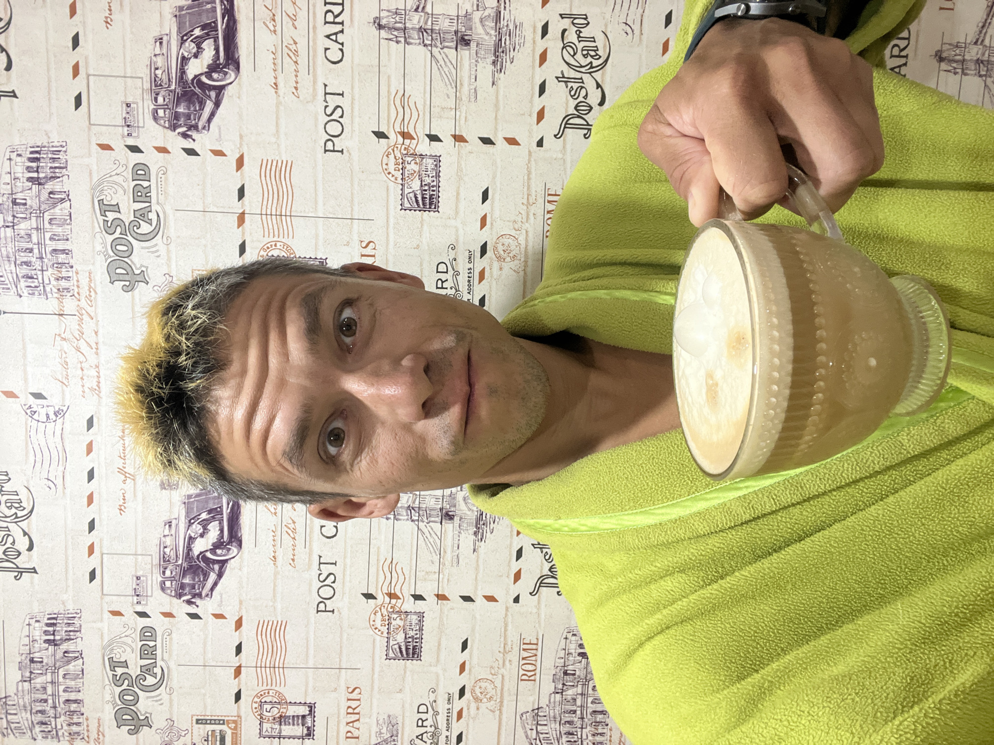 A man with blond-tipped hair, wearing a lime green bathrobe, holds a clear glass mug filled with a frothy beverage. He is looking directly at the camera with wide eyes and a slightly open mouth. The background is a patterned wallpaper depicting vintage postcards with images of the Colosseum, an old car, and the Tower Bridge, along with text such as "POST CARD" and "PARIS." The image was taken in Chaikovsky, Russia.